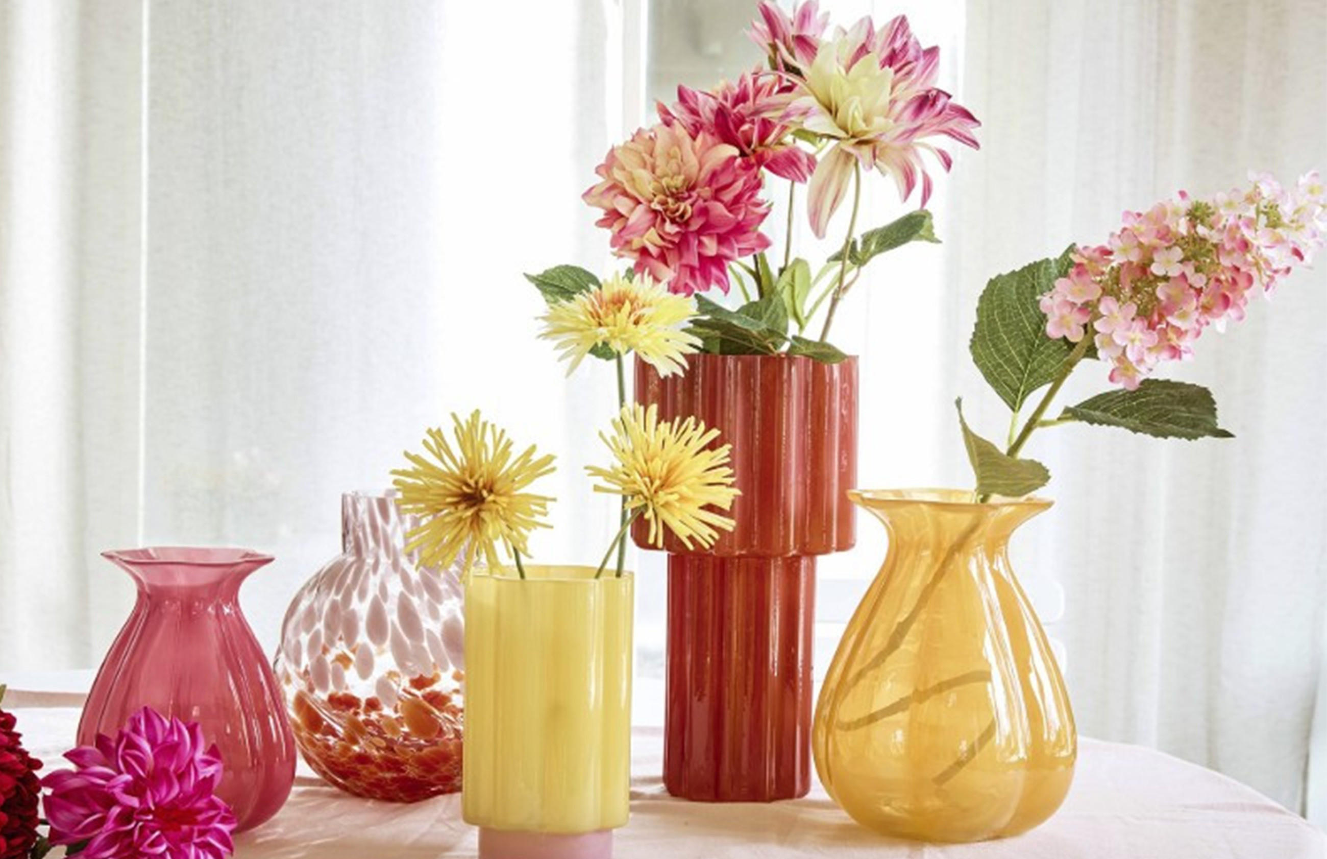 Mehrere unterschiedlich geformte und farbige Vasen mit verschiedenen Blumen auf einem weißen Tuch vor hellem Hintergrund.