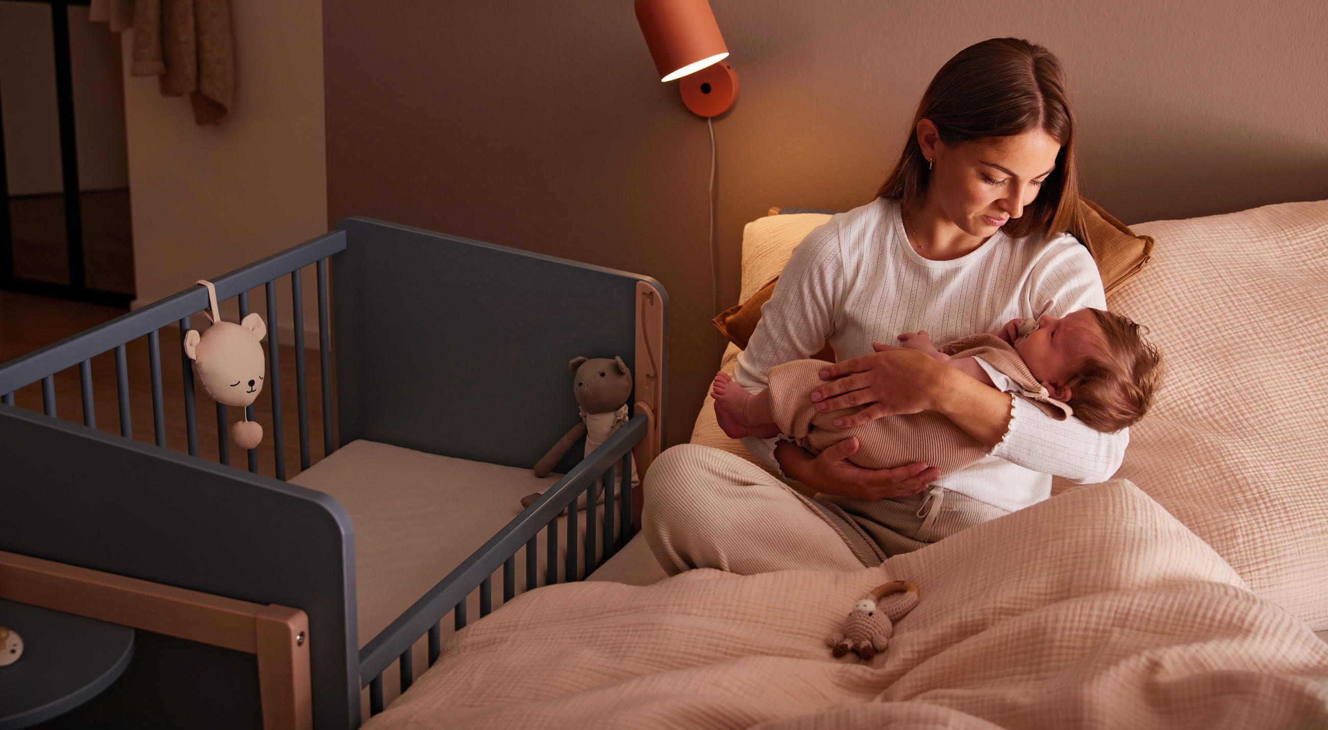 Eine Person sitzt auf einem Bett und hält ein Baby, daneben steht ein blaues Kinderbett mit zwei Stofftieren; die Szene ist in warmes Licht getaucht und wirkt ruhig und geborgen