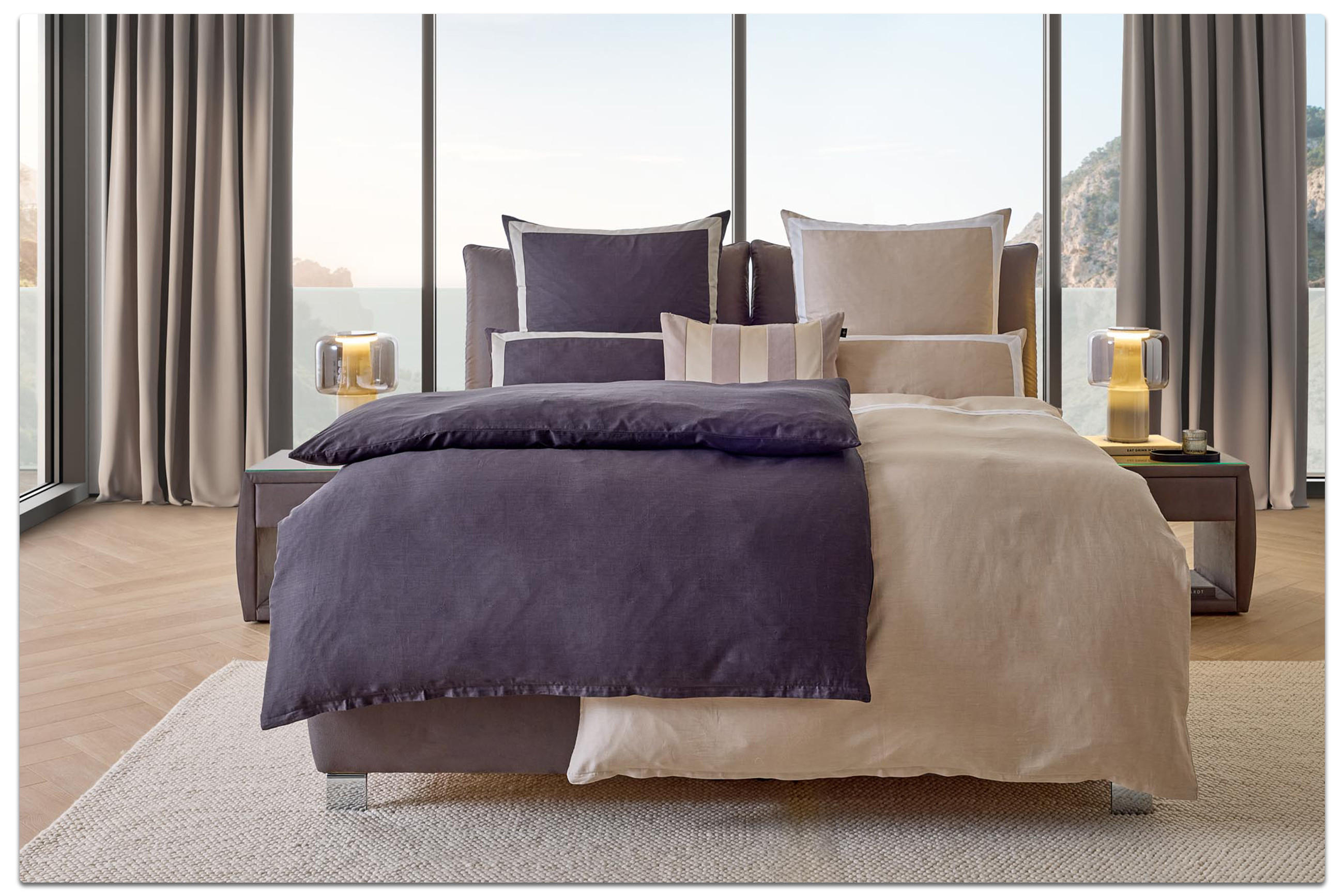 Luxuriöse Bettwäsche aus der Marcel Remus Kollektion in elegantem Dunkelviolett und Beige, dekoriert mit passenden Kissen auf einem modernen Doppelbett vor Panoramafenstern mit Meerblick.
