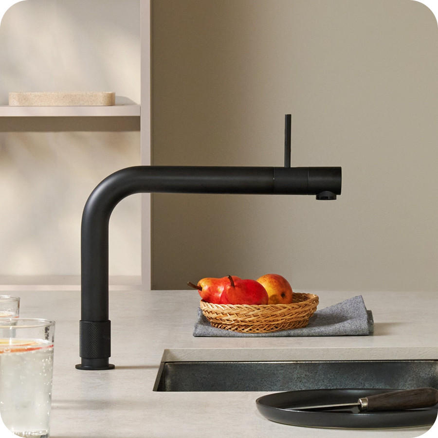 QUOOKER Küchenarmatur „FRONT“ in mattschwarzer Optik mit langem, horizontalem Auslauf über einer Spüle; auf der Arbeitsfläche stehen ein Glas Wasser sowie ein Korb mit Obst auf einem Tuch, im Vordergrund liegt ein Tablett mit Besteck.