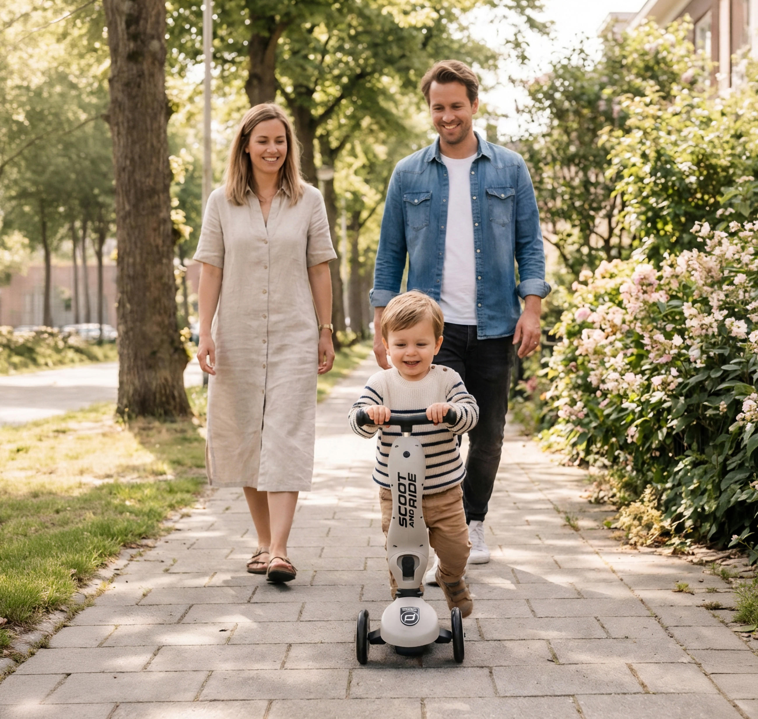 Kind auf einem Roller auf einem Gehweg zwischen Büschen und Bäumen mit zwei Erwachsenen, die dahinter gehen.