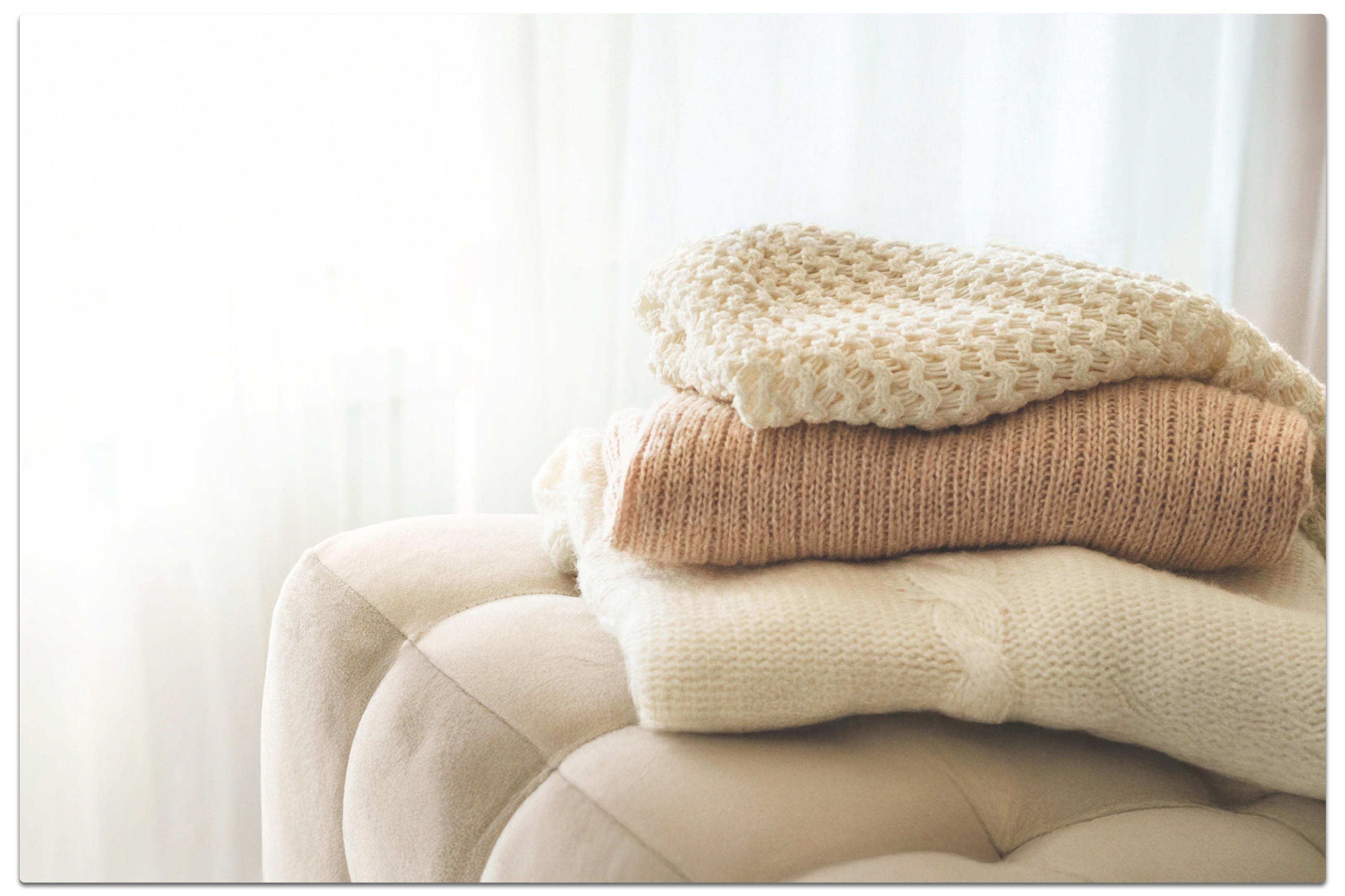 Ein weicher, beiger Hocker, auf dem drei gefaltete, helle Strickpullover in warmen Naturtönen liegen; im Hintergrund fällt weiches Tageslicht durch einen weißen Vorhang.