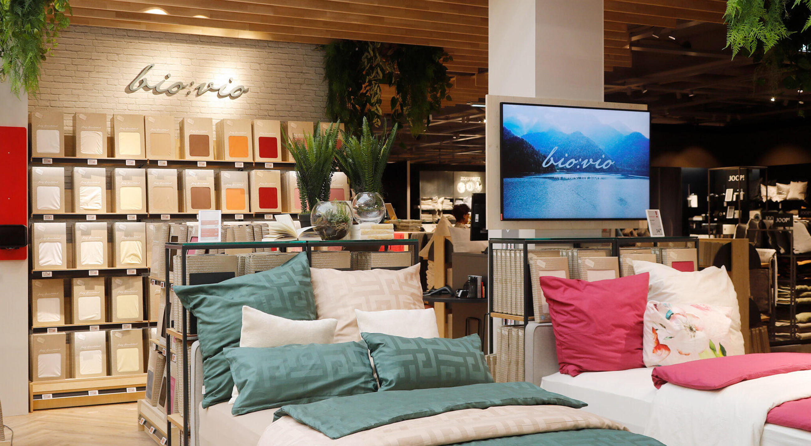 Ausstellung in einer XXXLutz-Filiale mit Bettwäsche und Kissen im Vordergrund, Regalwand mit Stoffmustern und „bio-vio“-Schriftzug sowie Bildschirm mit „bio-vio“-Motiv im Hintergrund.