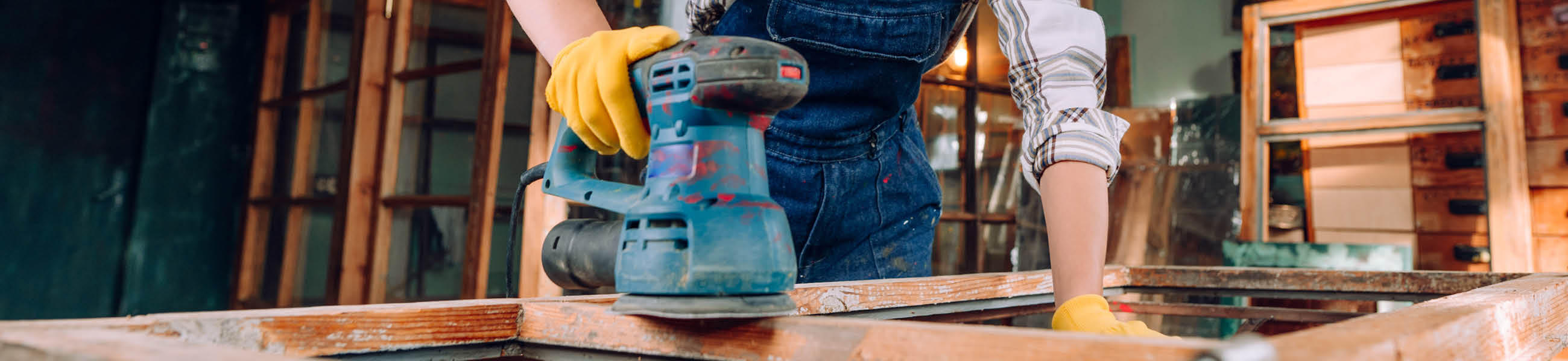 Person mit gelben Handschuhen, die einen blauen Schwingschleifer auf einem Holzstück verwendet, im Hintergrund Holzregale.