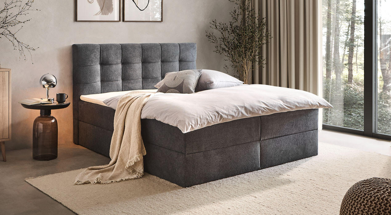 Dunkelgraues Boxspringbett mit Schlaftextilien, Nachttisch und Lampe gemütlich eingerichtet
