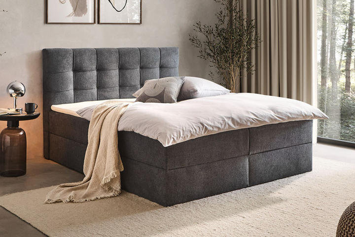 Dunkelgraues Boxspringbett mit Schlaftextilien, Nachttisch und Lampe gemütlich eingerichtet