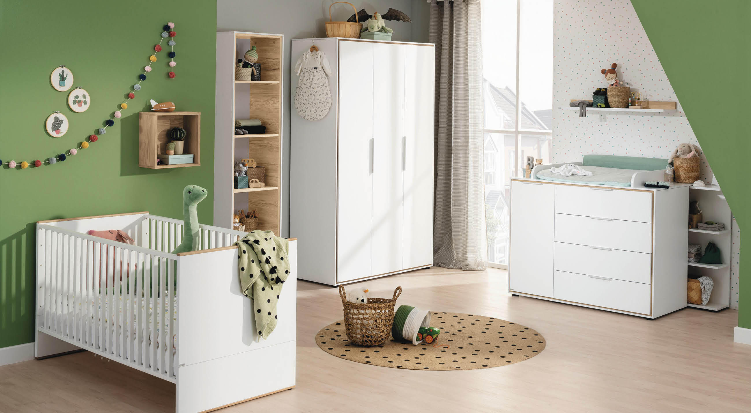 Kinderzimmer mit weißem Gitterbett, Kommode mit Wickelauflage, Spielzeugkorb und grüner Wanddekoration.