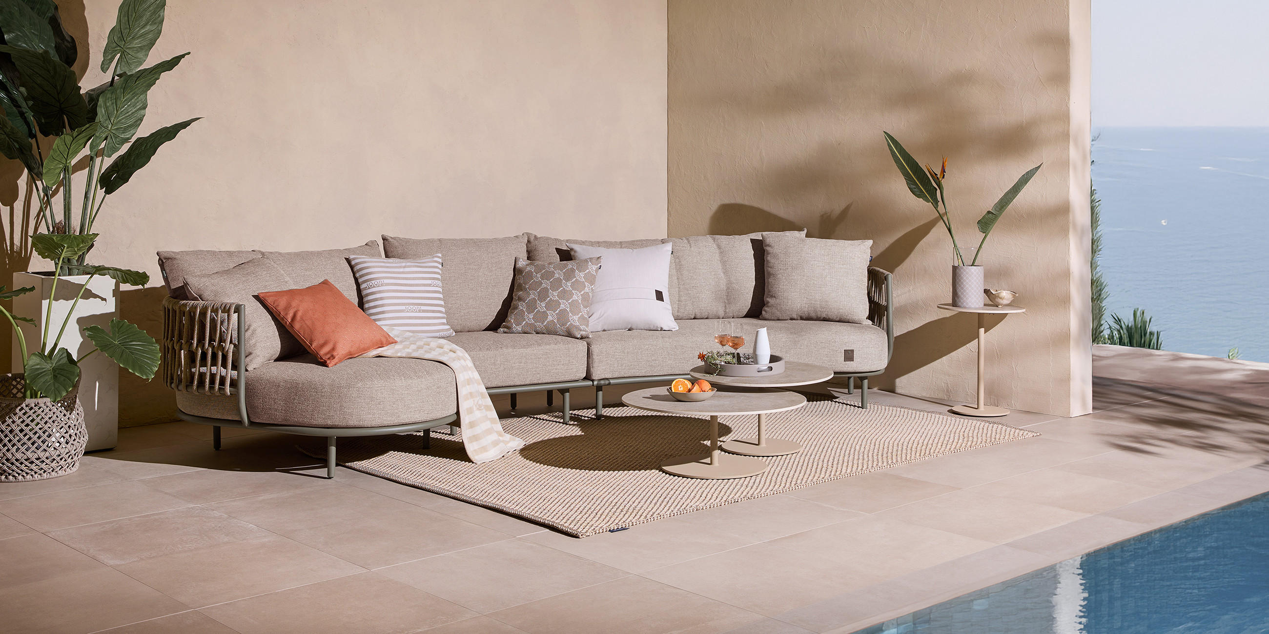 Outdoor Möbel und Textilien von JOOP! Living auf einer hellen Dachterrasse mit Sonnenschein und dekorativ platzierten Pflanzen, direkt am Wasser.