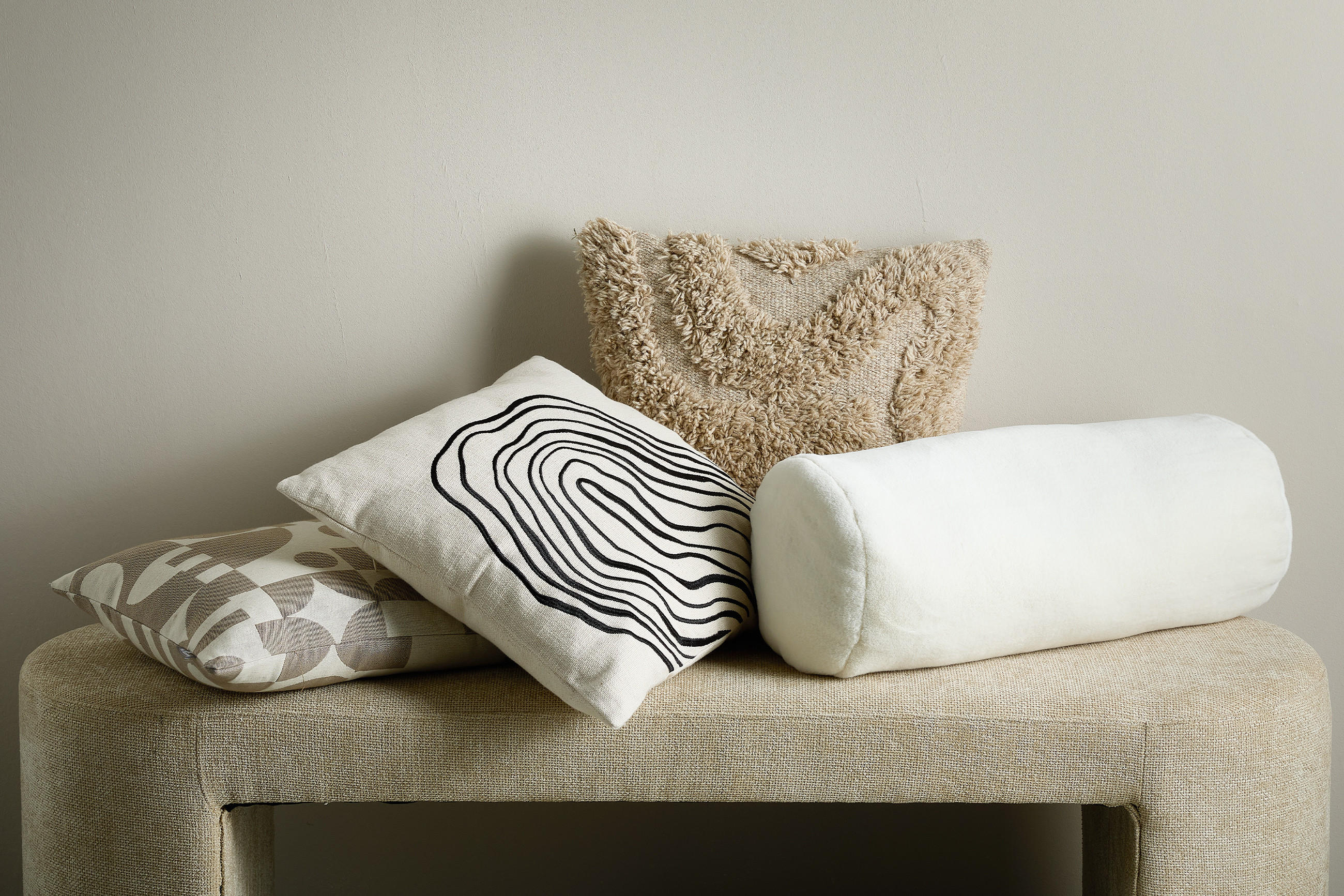 Vier dekorative Kissen in verschiedenen Formen und Texturen liegen auf einer hellen, gepolsterten Bank vor einer neutralen Wand.
