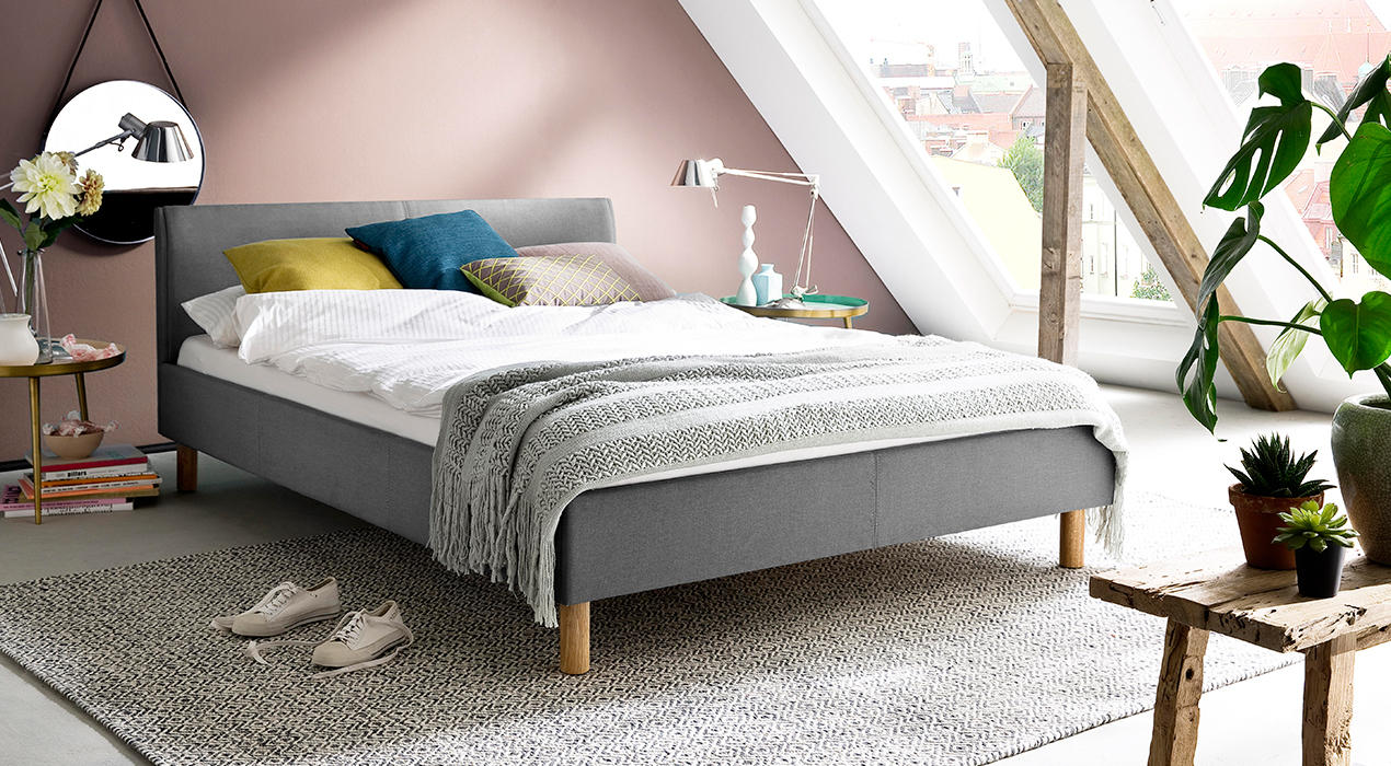 Graues Polsterbett mit Holzfüßen, Kissen und Decken.