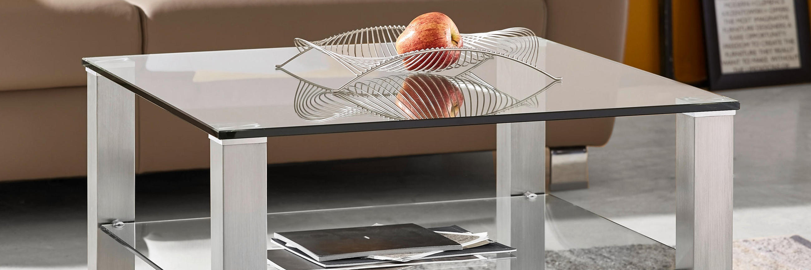 Quadratischer Couchtisch mit Glasplatte und Unterboden aus Glas, silbernen Metallbeinen und dekorativer Drahtschale mit Apfel steht auf gemustertem Teppich vor braunem Sofa