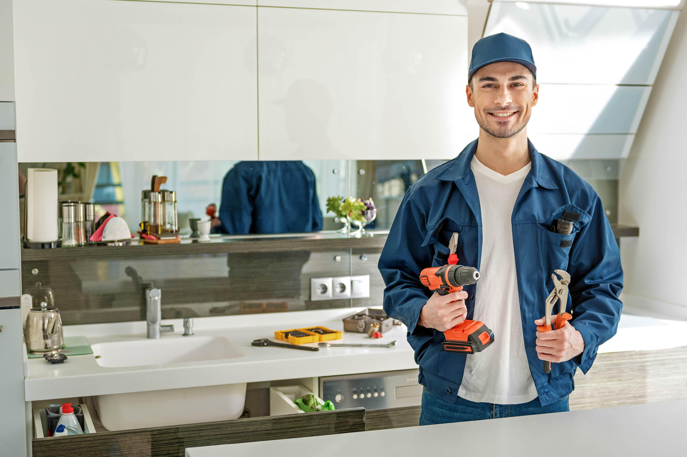 Junger Handwerker in blauer Arbeitskleidung hält eine Bohrmaschine und eine Zange in einer modernen Küche.