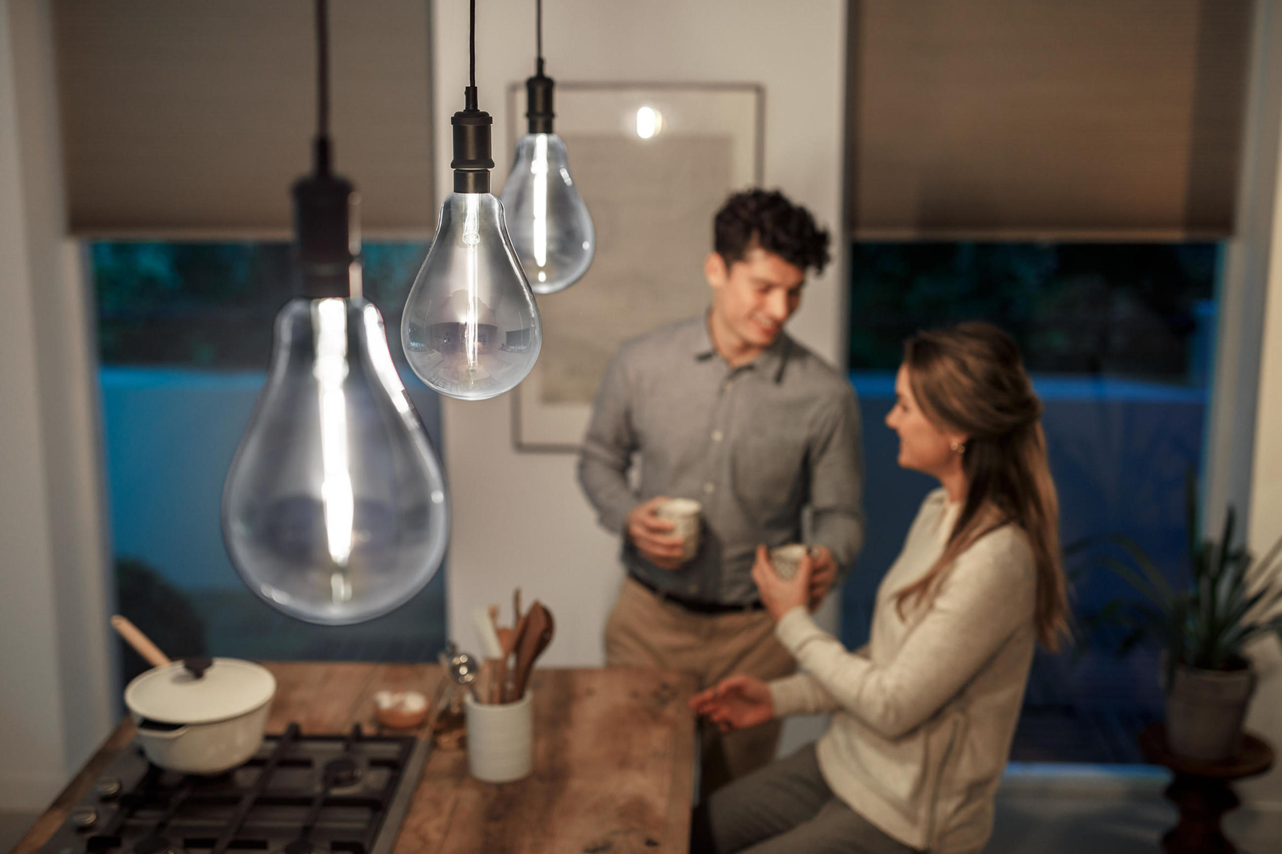 Nahaufnahme mehrerer hängender, transparenter PHILIPS Filament-Leuchtmittel über einer Kücheninsel; im Hintergrund unterhalten sich zwei Personen bei Getränken in stimmungsvoller Abendbeleuchtung.
