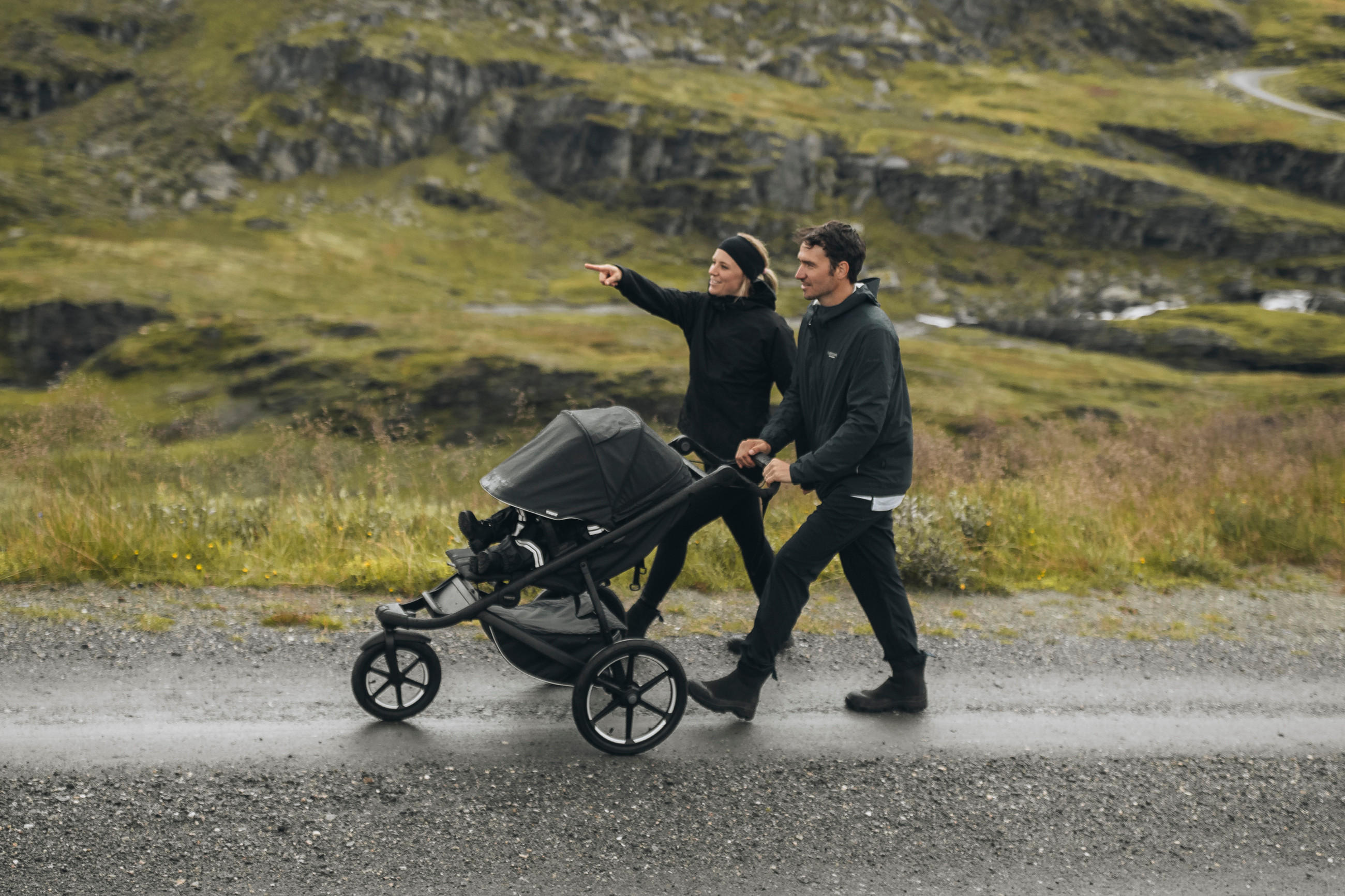 Paar in Outdoor-Kleidung schiebt einen schwarzen Thule Kinderwagen auf einem Weg in einer bergigen Landschaft