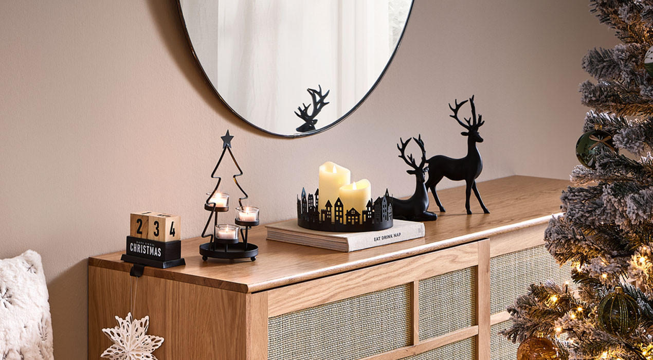 Gemütliche, weihnachtlich dekorierte Zimmerecke mit Holzschrank, Kerzen, Rentierfiguren, Spiegel, Sessel mit Kissen und beleuchtetem Weihnachtsbaum.