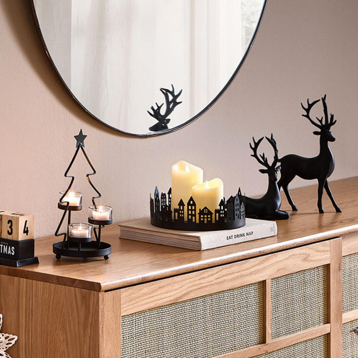 Gemütliche, weihnachtlich dekorierte Zimmerecke mit Holzschrank, Kerzen, Rentierfiguren, Spiegel, Sessel mit Kissen und beleuchtetem Weihnachtsbaum.