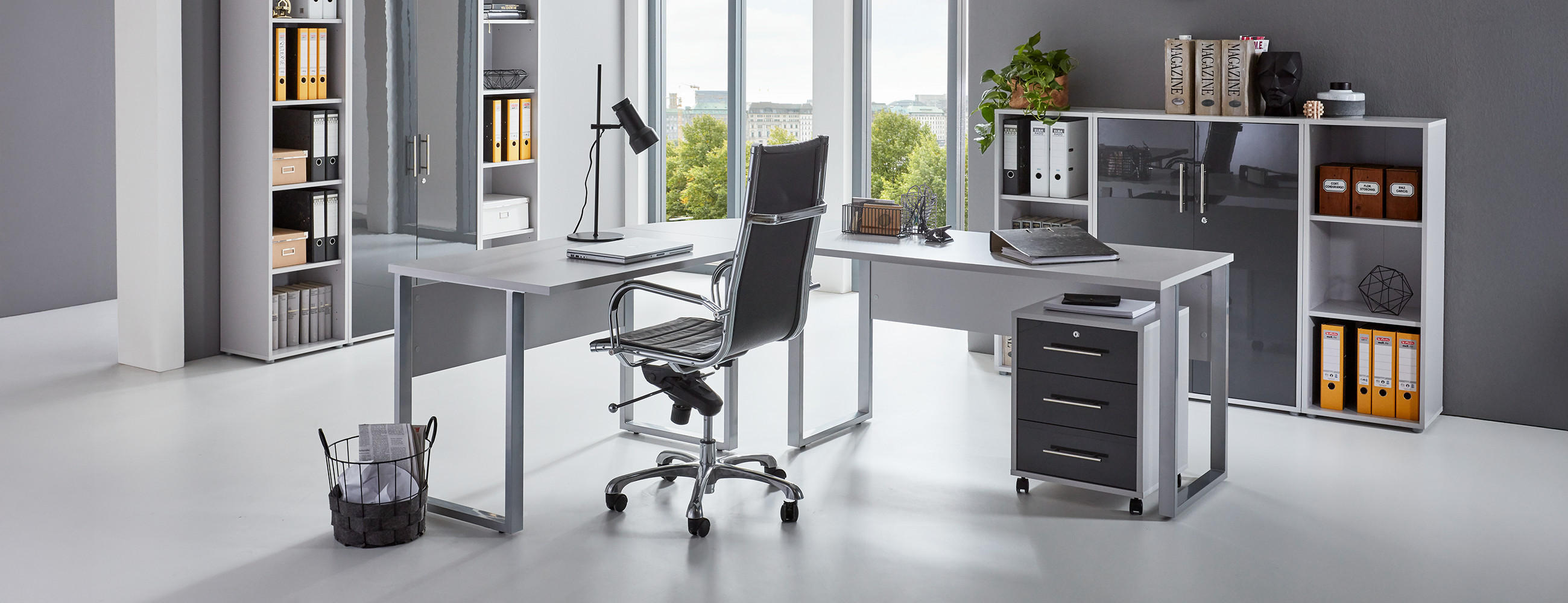 Modernes Büro mit großem Schreibtisch, Laptop, Lampe und Dokumenten, umgeben von Regalen und Aktenschränken; im Hintergrund große Fenster mit Blick ins Grüne.