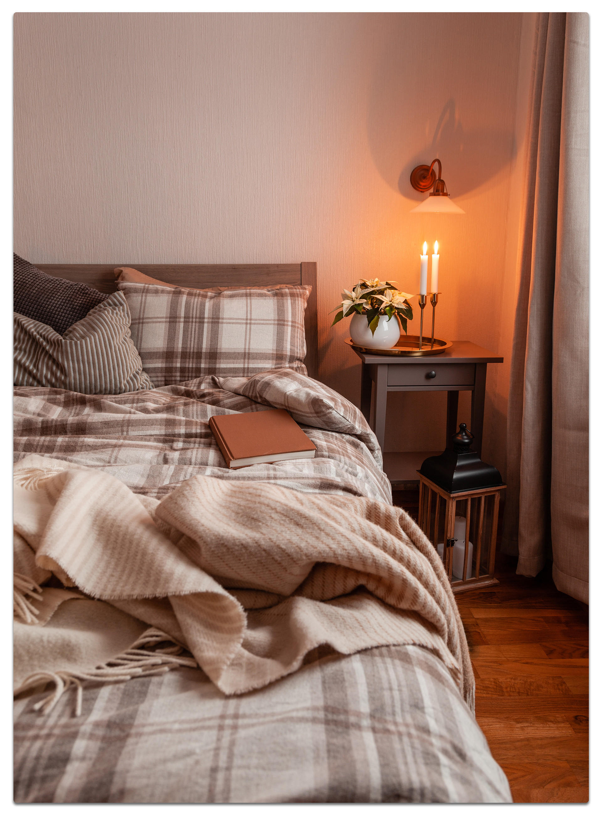 Gemütliches Schlafzimmer mit kariertem Bettbezug, beiger Decke, Buch und warmem Kerzenlicht auf dem Nachttisch – behagliches Cocooning-Ambiente mit natürlichen Materialien und sanfter Beleuchtung.