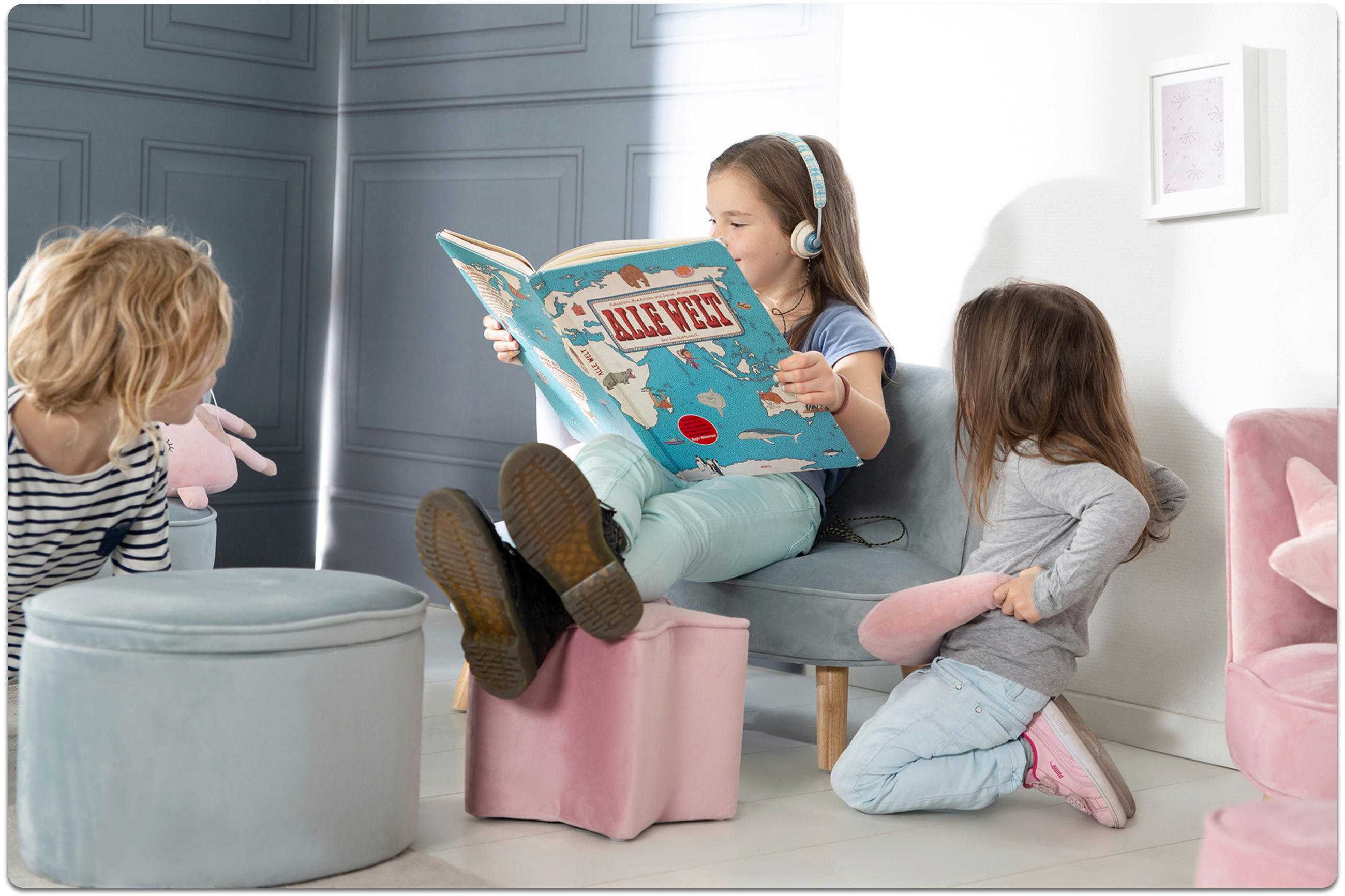Drei Kinder sitzen in einem hellen Zimmer, eines liest ein großes Buch mit Kopfhörern, die anderen beiden schauen zu.