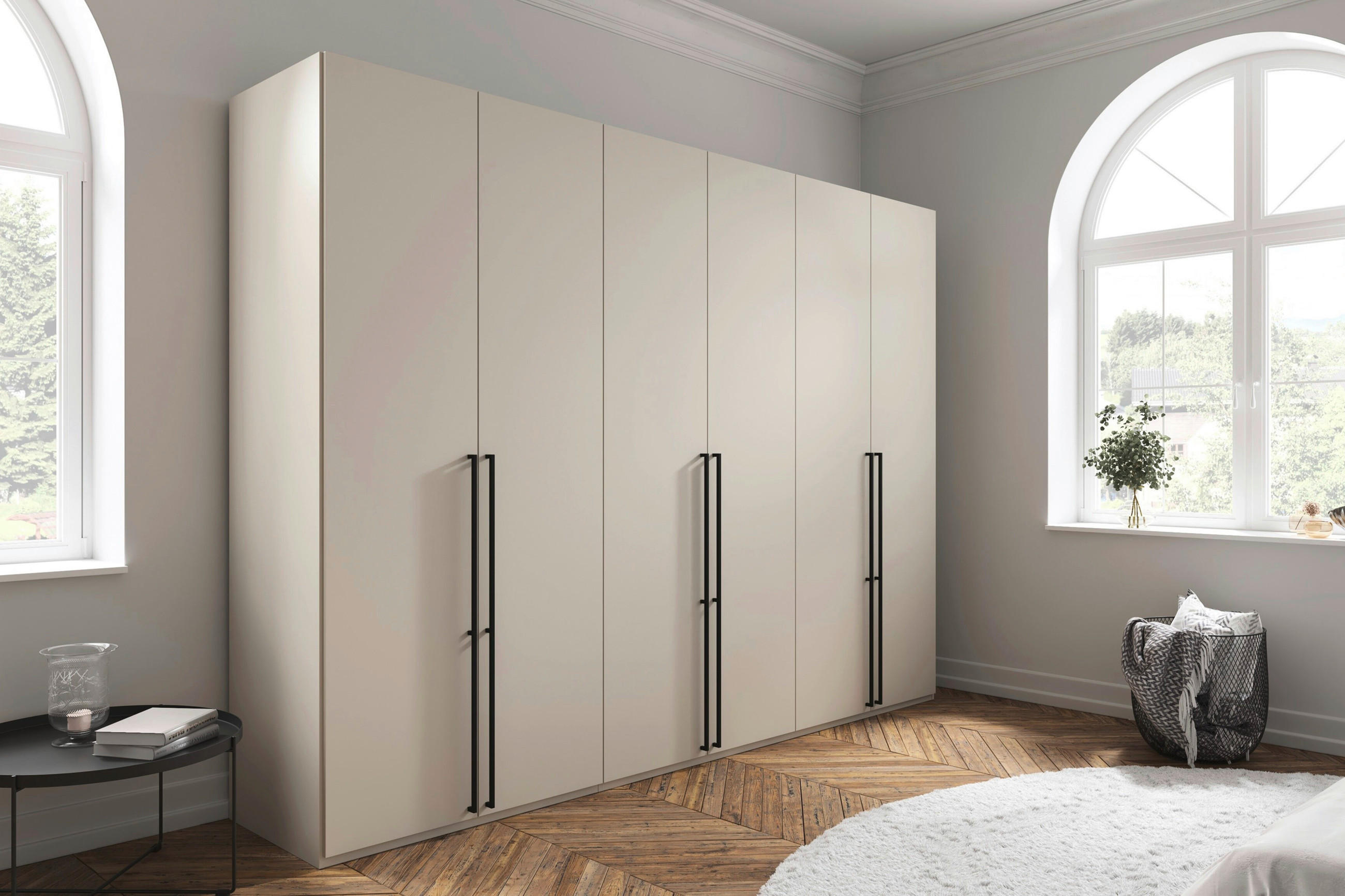 Heller Kleiderschrank mit mehreren Türen und schwarzen Griffen in lichtdurchflutetem Zimmer mit großen Rundbogenfenstern und Holzparkett.