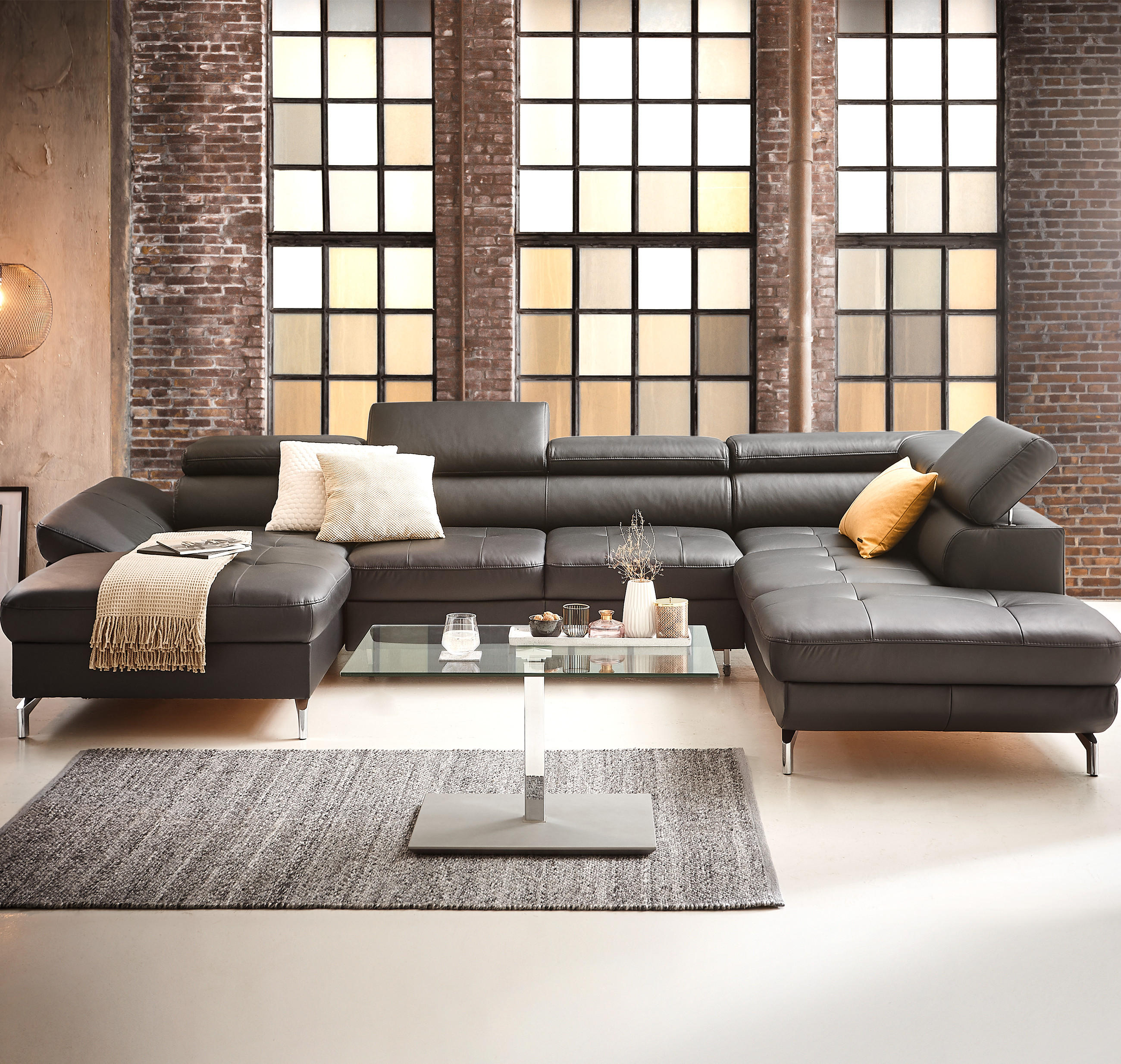Großes, modernes Ecksofa mit Kopfstützen, Kissen und Decke vor Backsteinwand und Glas-Couchtisch.