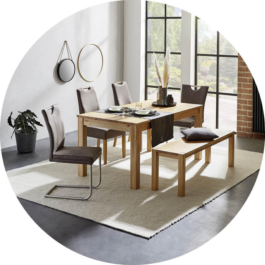 Esszimmer mit hellem Holztisch, vier gepolsterten Stühlen, einer Holzbank mit Kissen, zwei runden Wandspiegeln und einer Pflanze auf hellem Teppich vor großen Fenstern