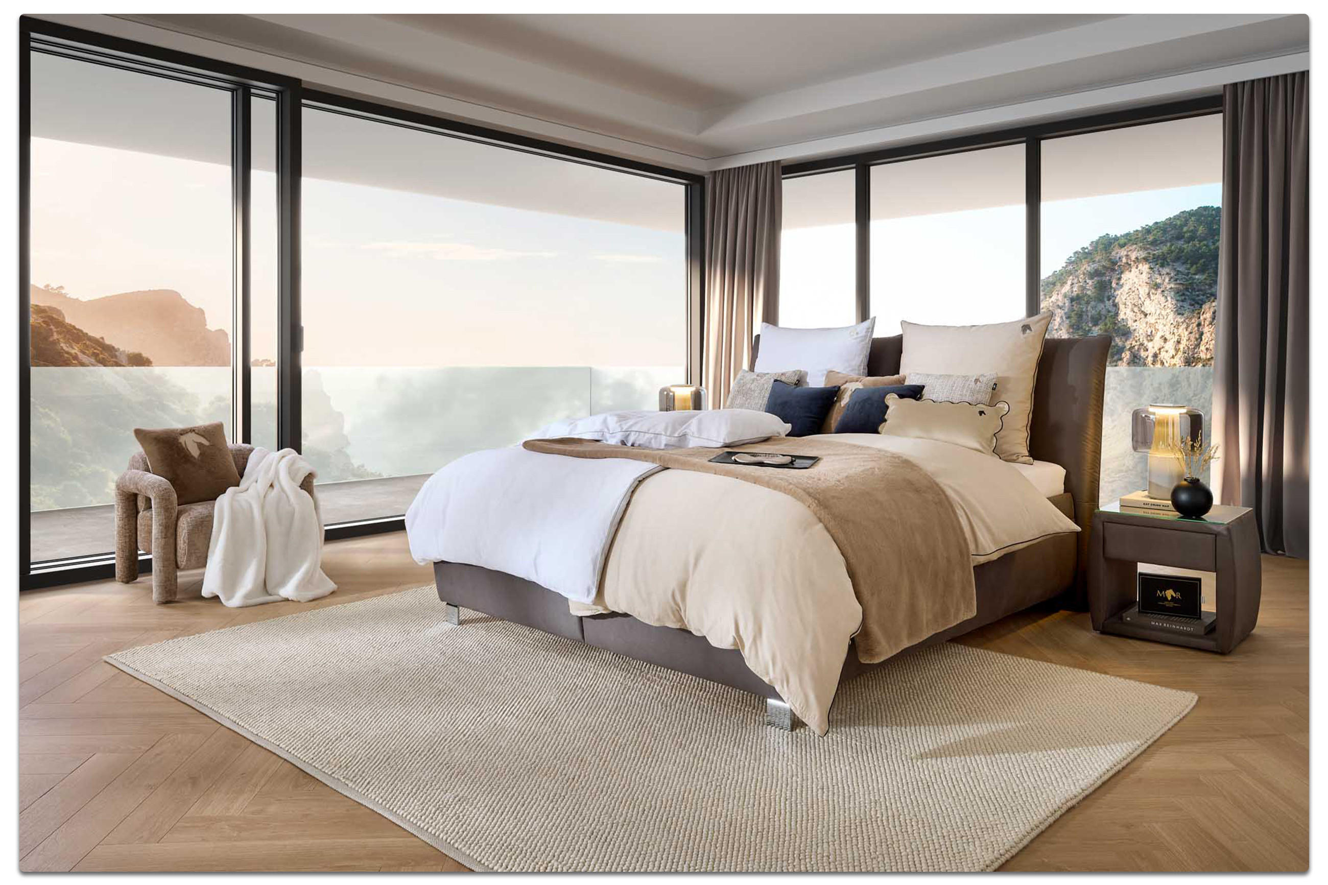 uxuriöse Bettwäsche aus der Marcel Remus Kollektion auf einem modernen Doppelbett mit neutralen Beige- und Brauntönen, dekoriert mit Kissen und Decke, vor Panoramafenstern mit Meerblick.