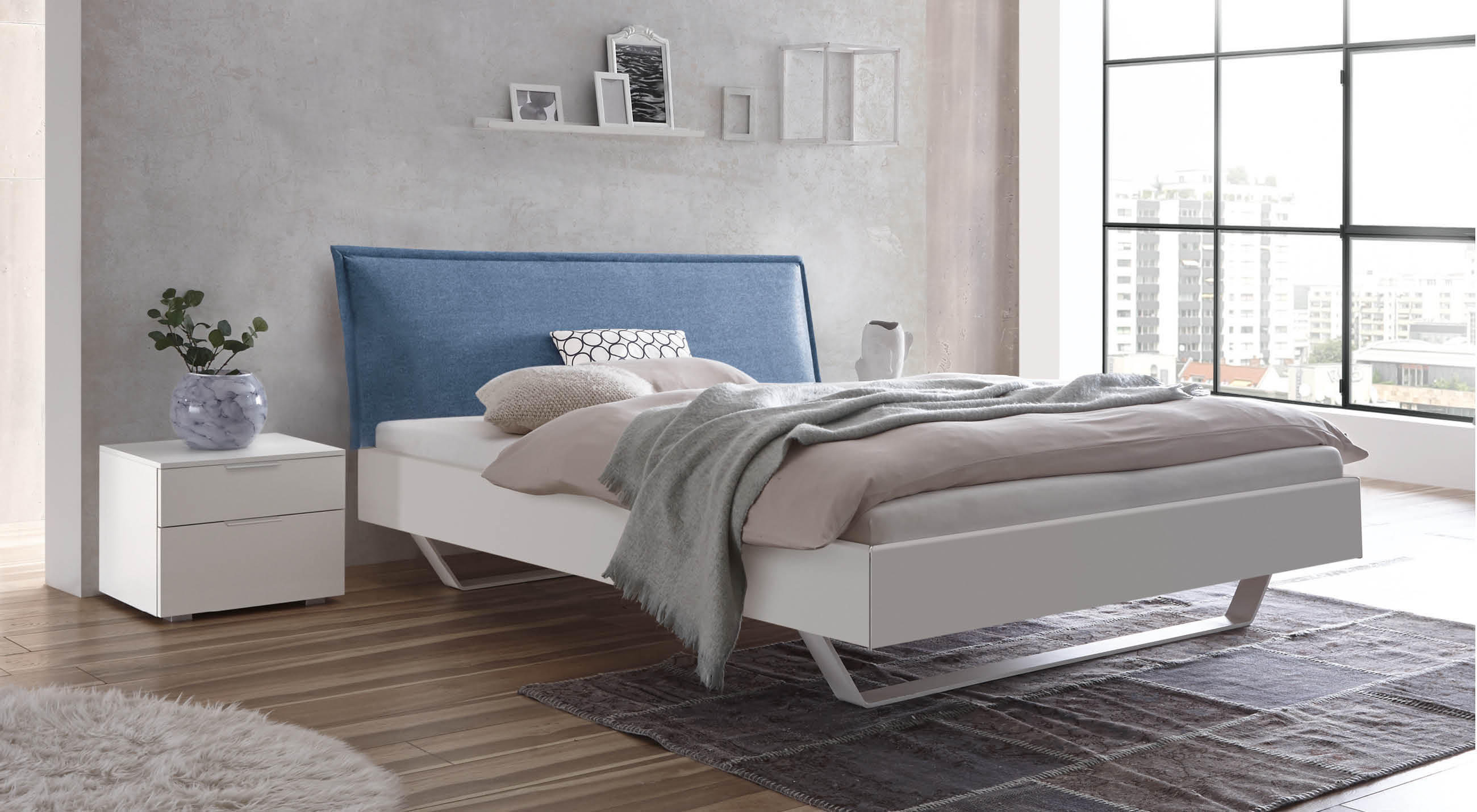 Modernes Hasena MDF-Bett in Weiß mit schrägem, blau gepolstertem Kopfteil in einem hellen Schlafzimmer mit großer Fensterfront und minimalistischen Möbeln.