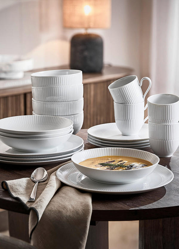 Bela porcelanasta posoda na okrogli leseni mizi z juho, krožniki, skodelicami in skledami v elegantni jedilnici.