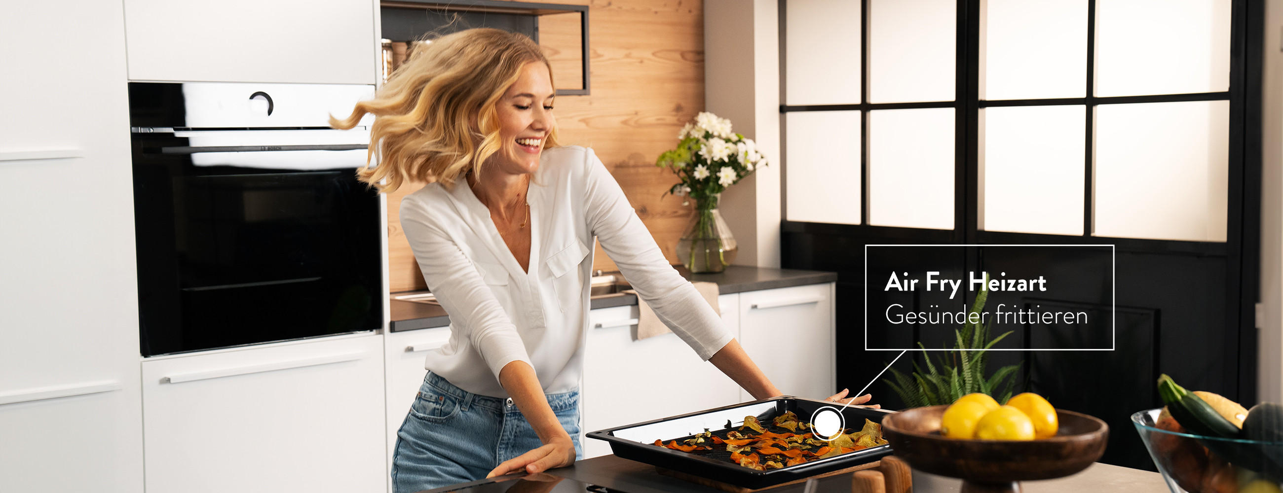 Backofen von BOSCH, blonde Frau in weißer Küche, Einbaub-Backofen von BOSCH - Der Einbau-Backofen mit digitalem Bedienring, TFT-Touchdisplay, Bosch Assist und Air Fry. Zusätzlich kann Home Connect bei der Bedienung über die App genutzt werden