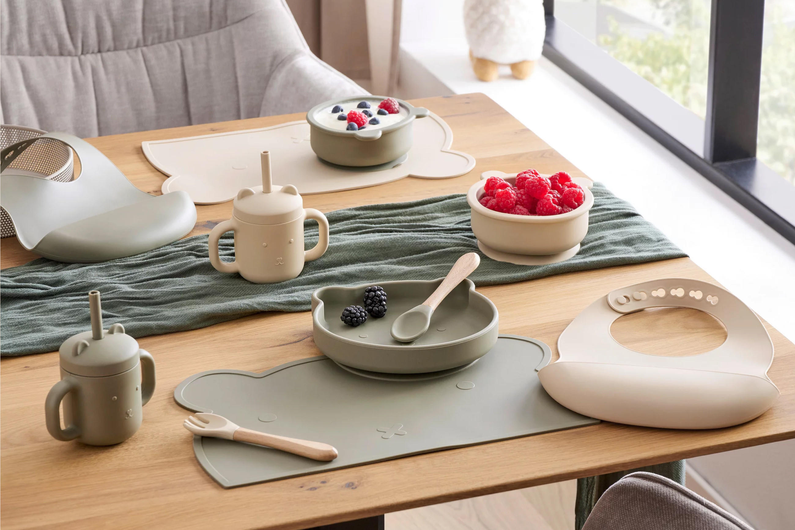 Kindergeschirr aus Silikon mit Tellern, Bechern, Löffel und Gabel auf einem Holztisch neben einem Fenster.