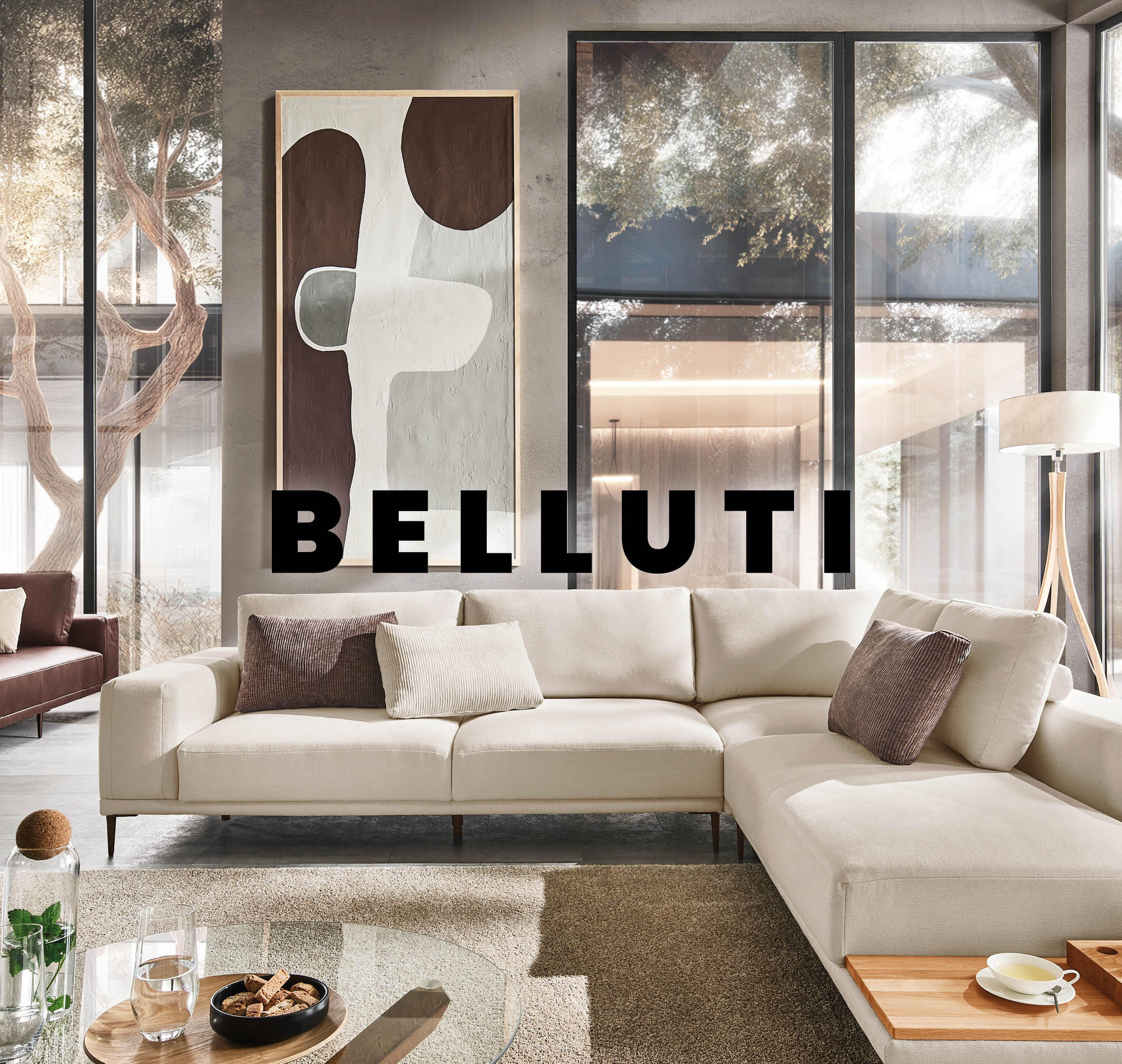 Modernes Wohnzimmer mit beigem Ecksofa und brauner Liege, Glasfronten mit Gartenblick, Couchtisch mit Teeservice und Gläsern, an der Wand abstraktes Bild, mittig Schriftzug ‚BELLUTI‘