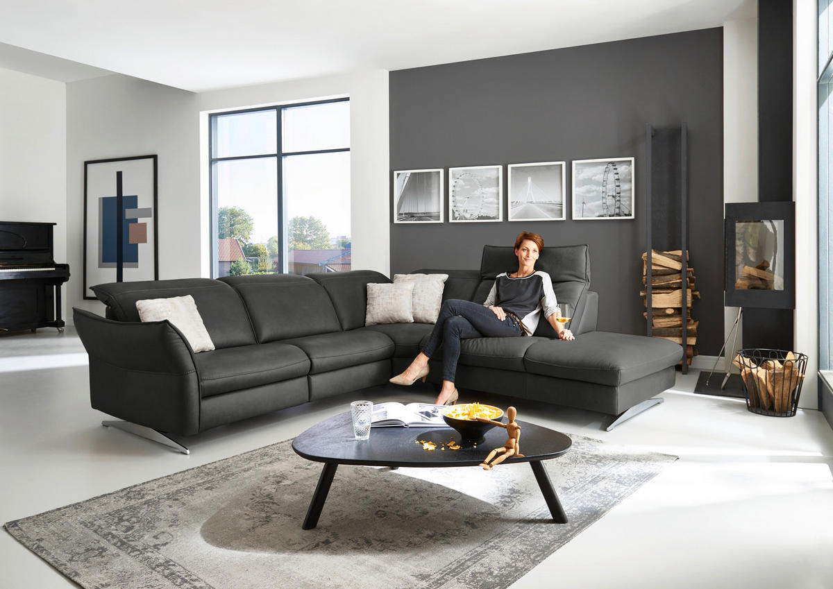 Hukla ECKSOFA inkl. Funktionen Valdera Schwarz Echtleder, links davon: Hochflorteppich, Kissen in weiß, rechts davon: Couchtisch, Kamin in hellem Wohnzimmer.