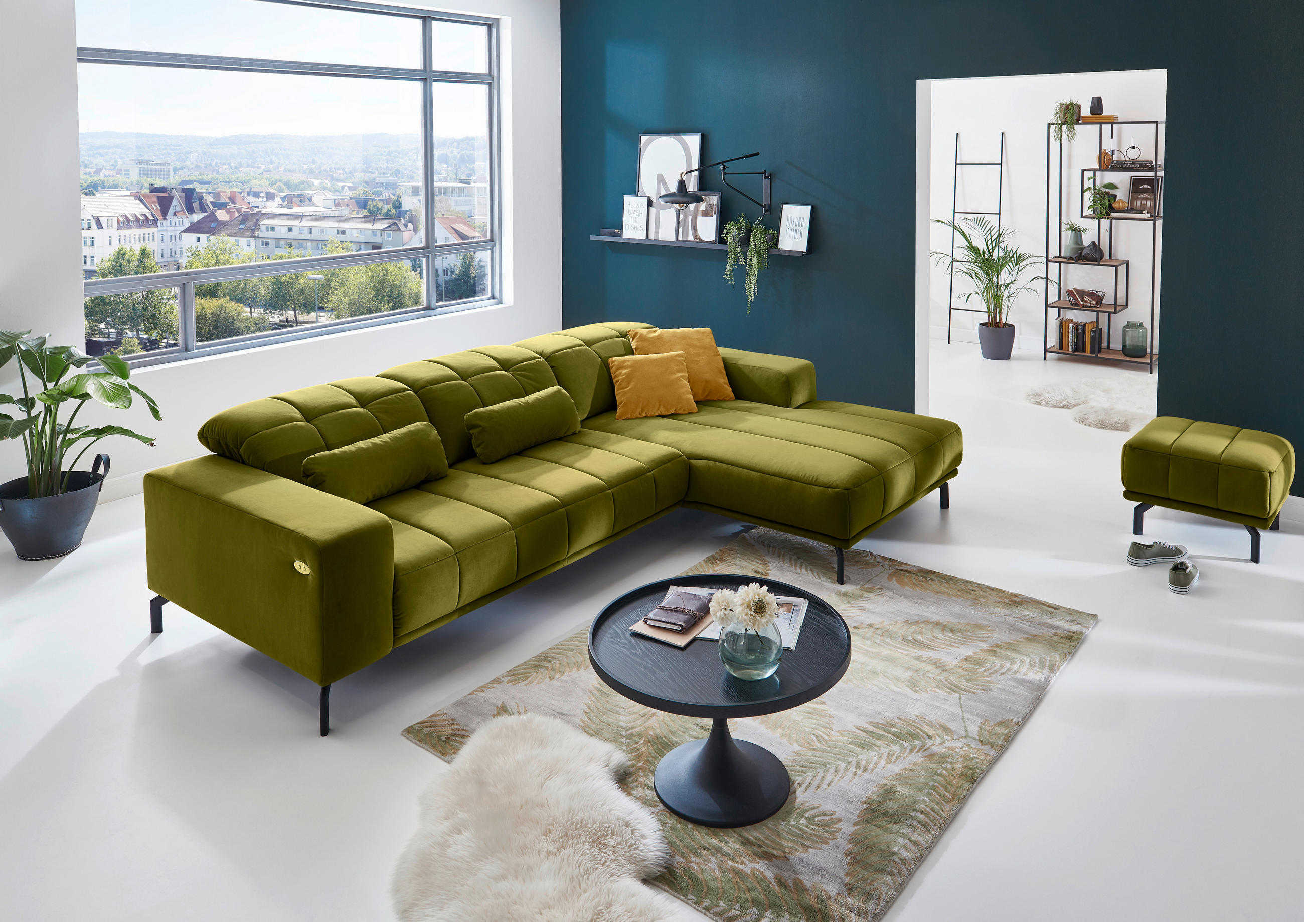 Beldomo Style ECKSOFA Grün Velours, links davon: Topfpflanze in grün, rechts davon: Kunstfell, Couchtisch, Lektüre in hellem Wohnzimmer.