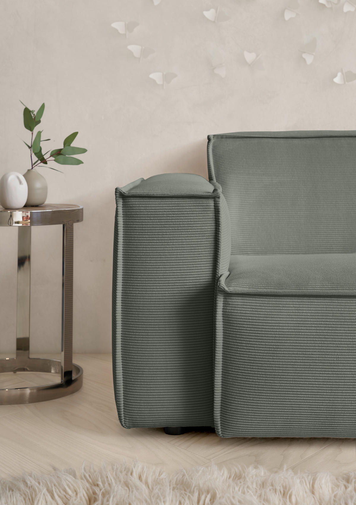 Hom`in Kunstfell, links davon: Blume in grün, Vase in weiß, rechts davon: Sofa, Decke in hellem Wohnzimmer.