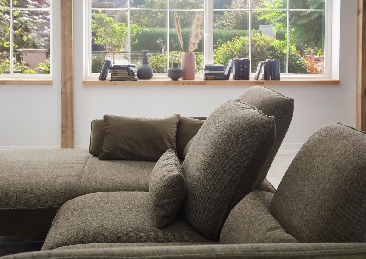 Moderano ECKSOFA Olivgrün Flachgewebe, rechts davon: Kissen, Lektüre in schwarz, Vase in hellem Wohnzimmer.