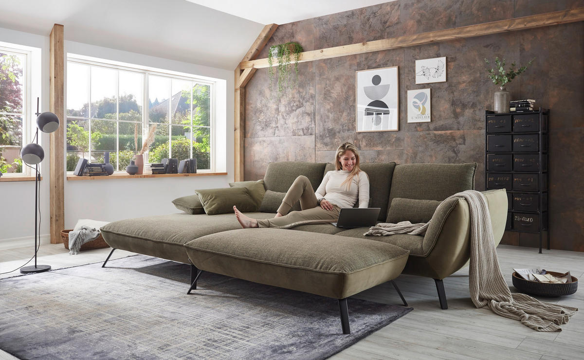 Moderano ECKSOFA Olivgrün Flachgewebe, links davon: Hochflorteppich, Kissen, rechts davon: Highboard, Lektüre in hellem Wohnzimmer.