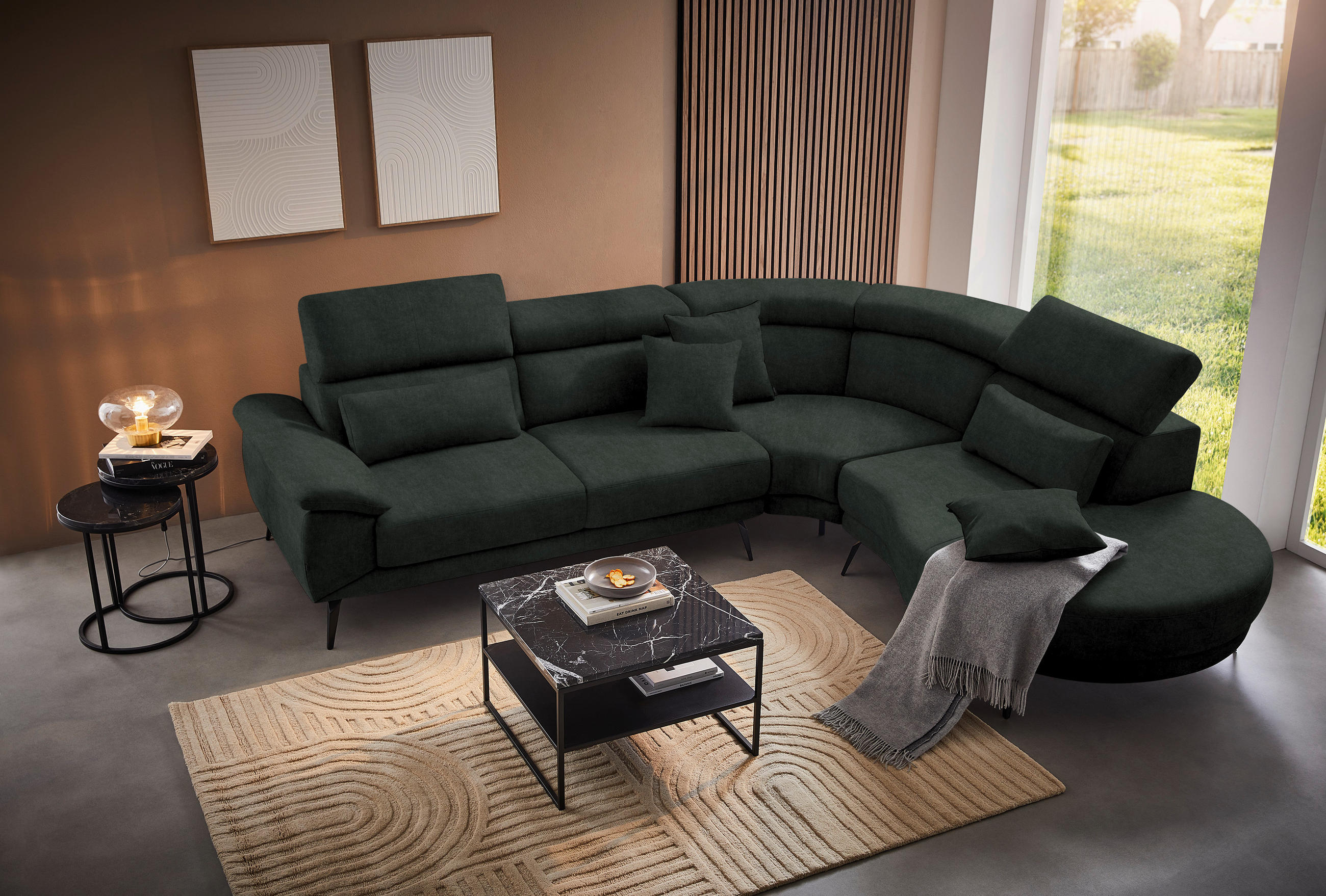 Novel ECKSOFA Dunkelgrün Velours, links davon: Webteppich, rechts davon: Decke in grau, Couchtisch, Kissen in grün in dunklem Wohnzimmer.
