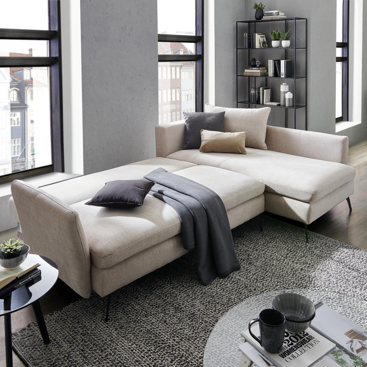 XXXL ECKSOFA Schwarz Webstoff, links davon: Webteppich in grau, Beistelltisch in schwarz, rechts davon: Decke, Regal in hellem Wohnzimmer.