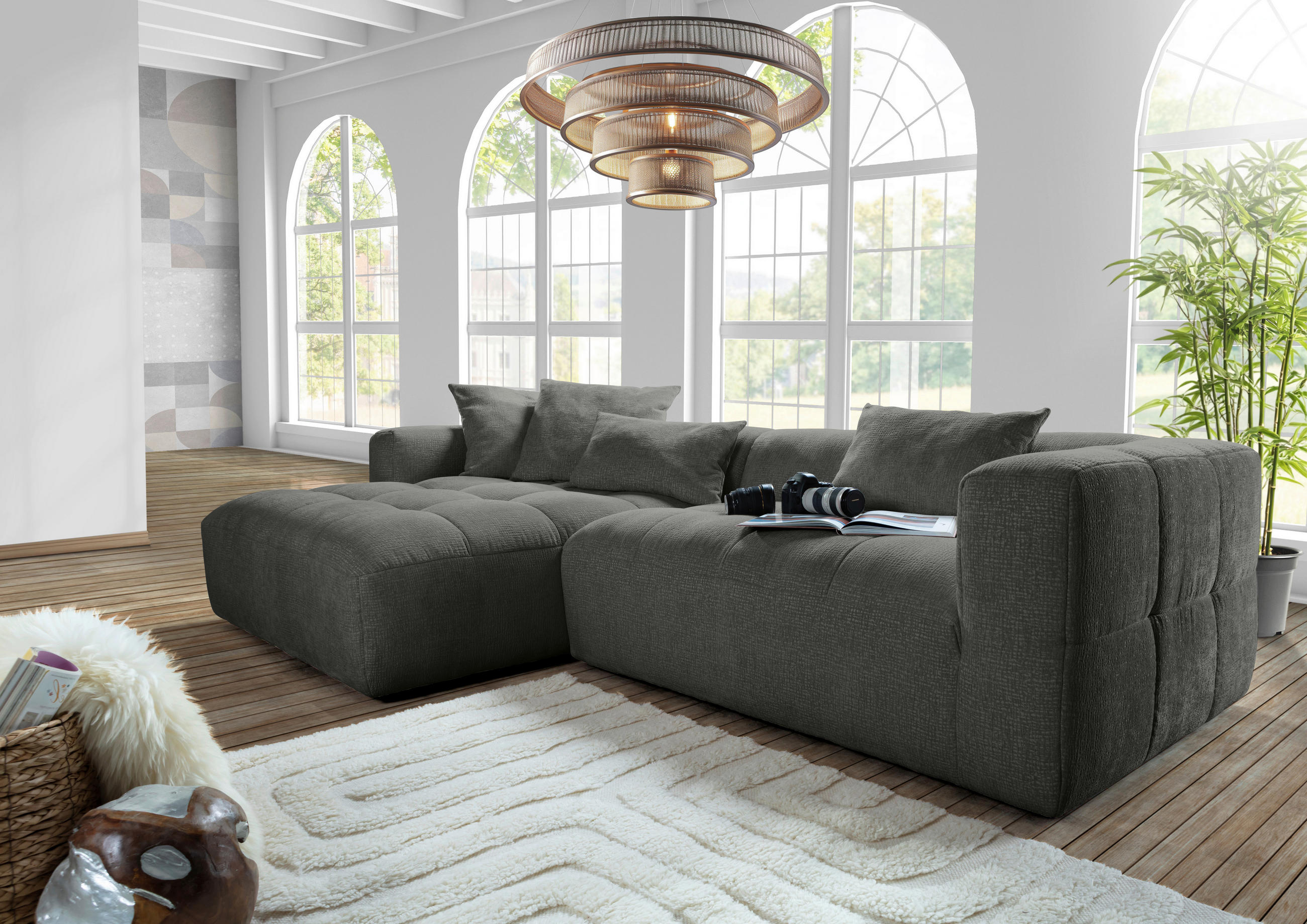 Carryhome ECKSOFA Schwarz Flachgewebe, links davon: Hochflorteppich in weiß, rechts davon: Deckenbeleuchtung, Topfpflanze in hellem Wohnzimmer.