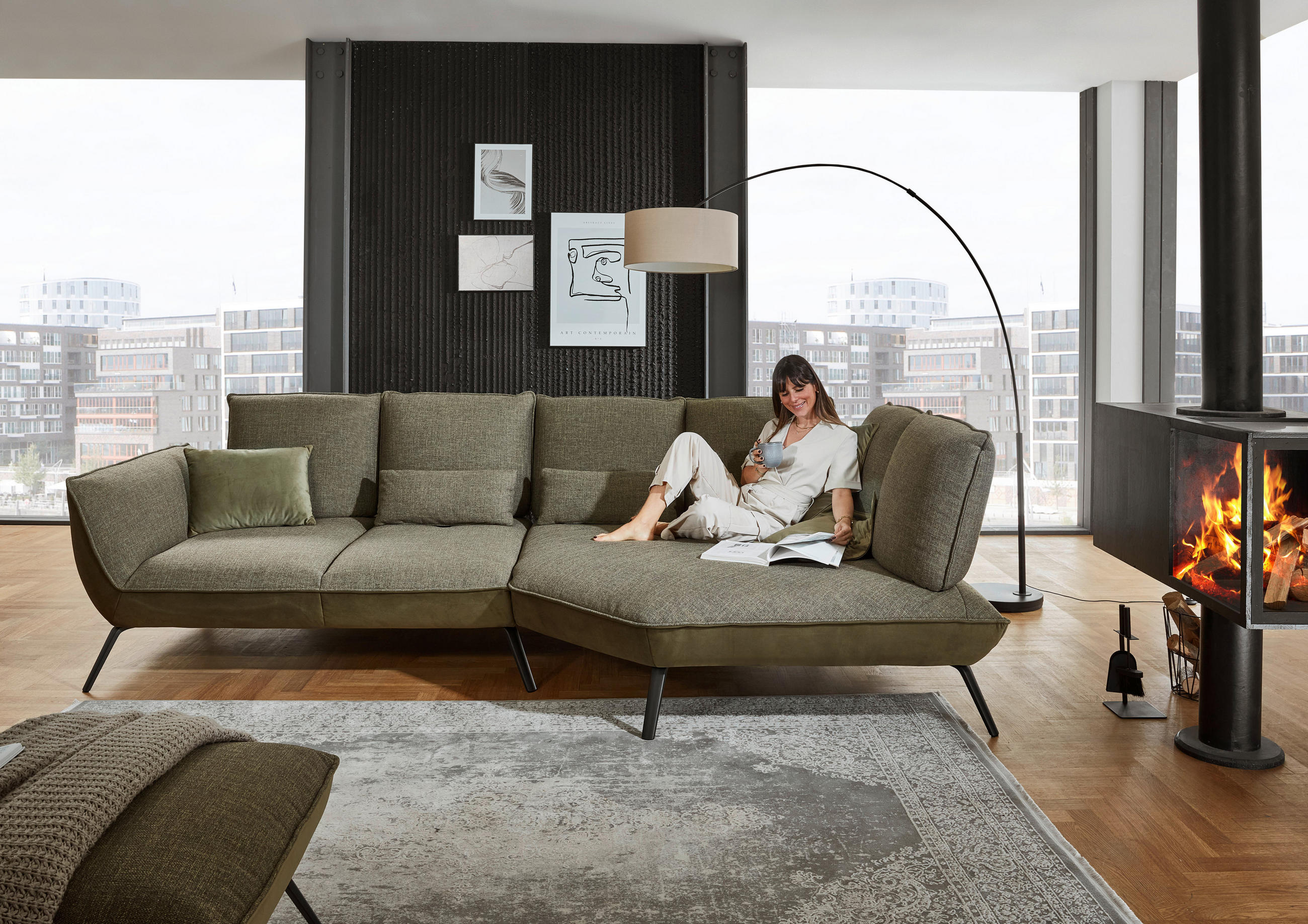Moderano ECKSOFA Olivgrün Flachgewebe, links davon: Kissen in grün, rechts davon: Hochflorteppich, Stehleuchte, Bild in weiß in hellem Wohnzimmer.