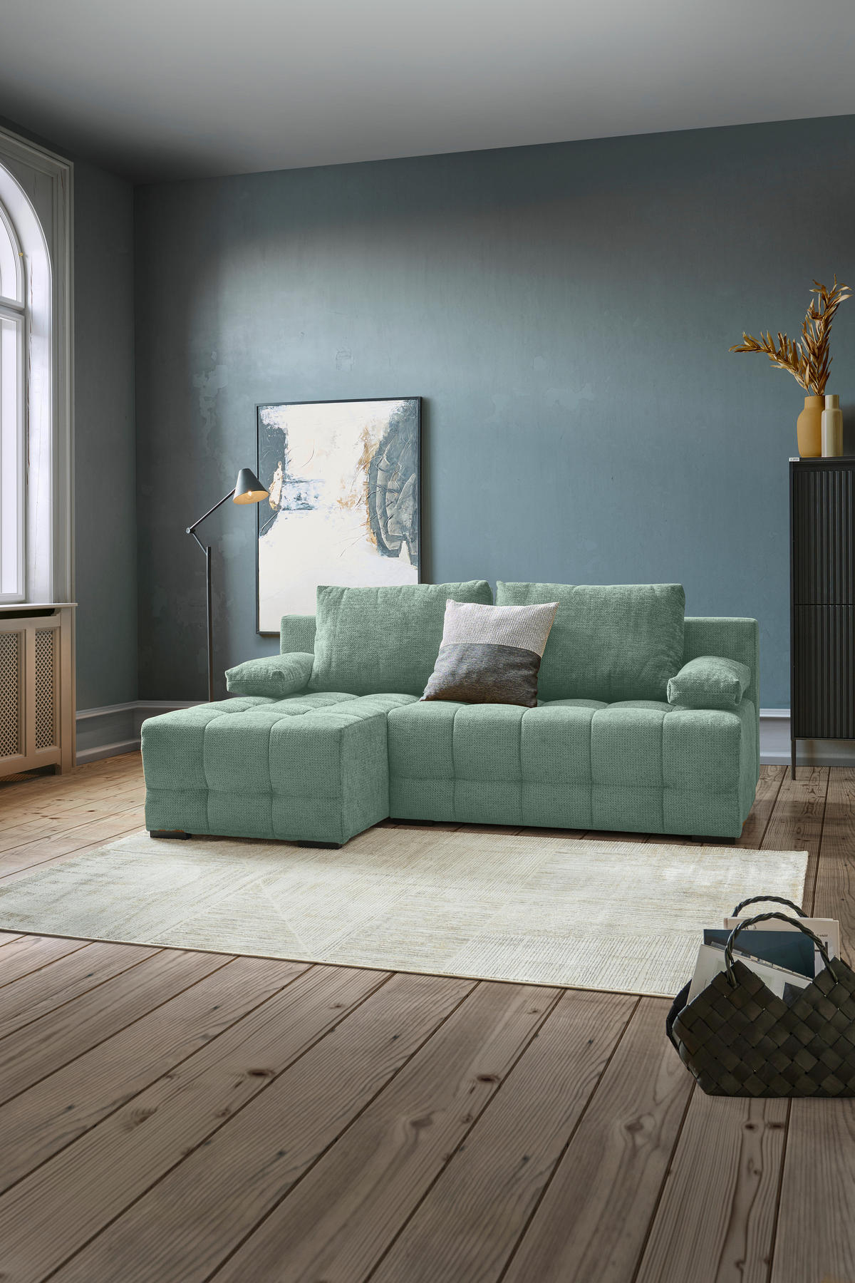home24 ECKSOFA Dunkelgrün Chenille, links davon: Hochflorteppich in grün, Boxspringbett in grün, Bild, Tischleuchte in dunklem Wohnzimmer.