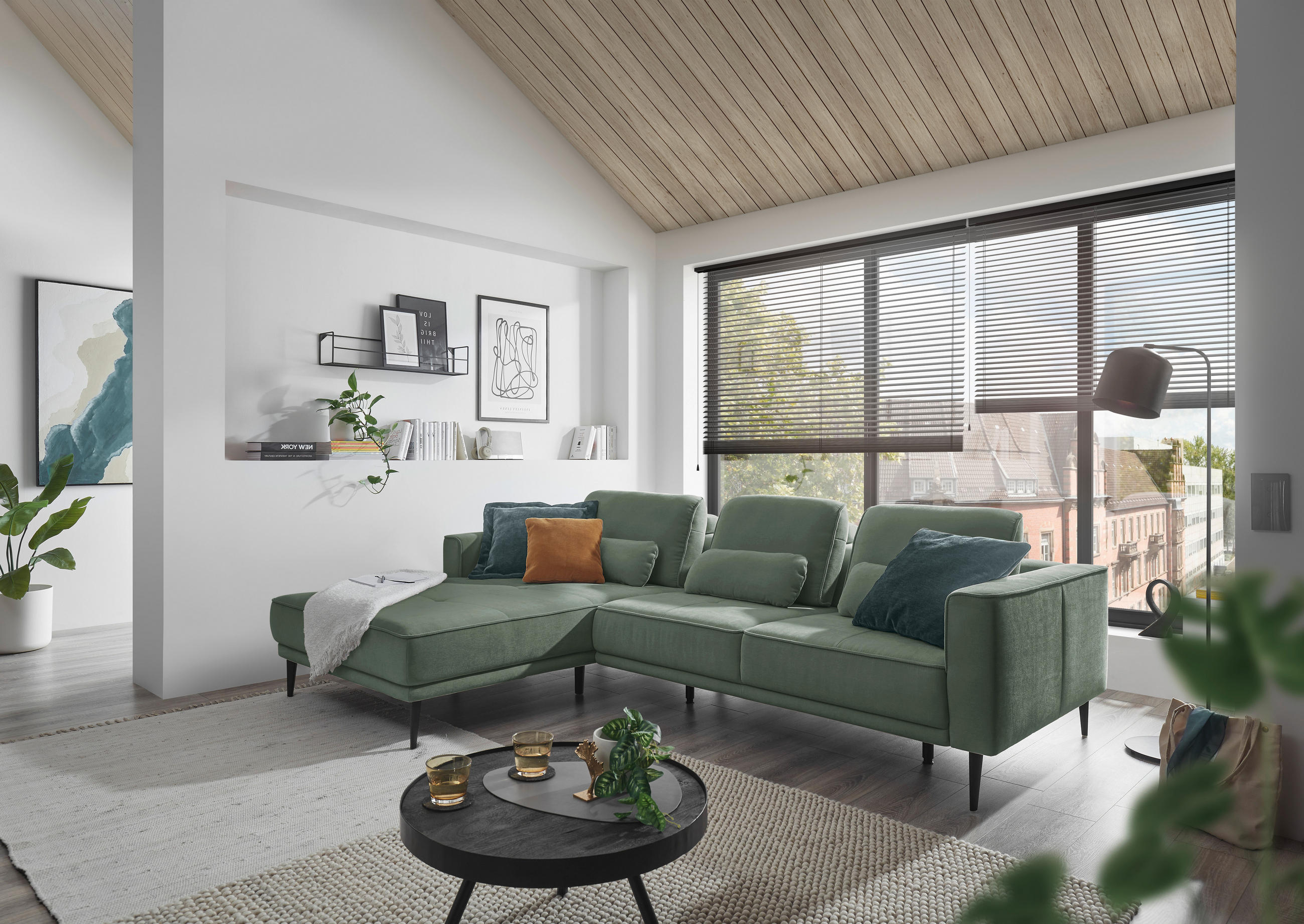 SetOne by Musterring ECKSOFA Dunkelgrün Flachgewebe, links davon: Webteppich, Couchtisch in schwarz, rechts davon: Plissee, Kissen in hellem Wohnzimmer.