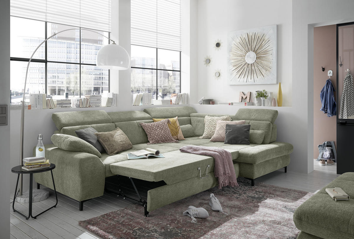 SetOne by Musterring ECKSOFA Grün Webstoff, rechts davon: Webteppich, Decke, Hocker in grün, Jalousie in weiß in hellem Wohnzimmer.