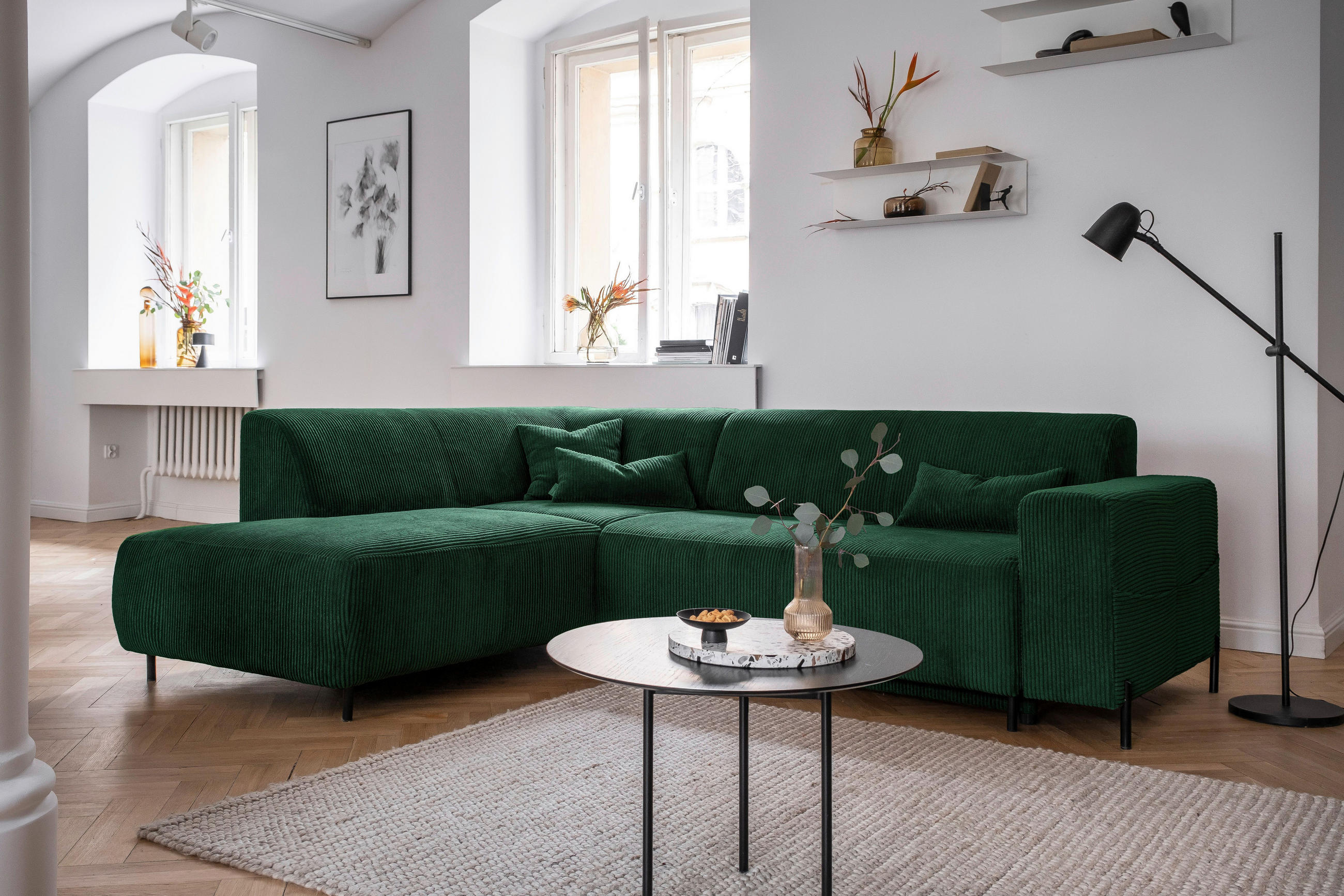 MID.YOU ECKSOFA Dunkelgrün Cord, rechts davon: Hochflorteppich, Couchtisch, Beistelltisch, Stehleuchte in grün in hellem Wohnzimmer.