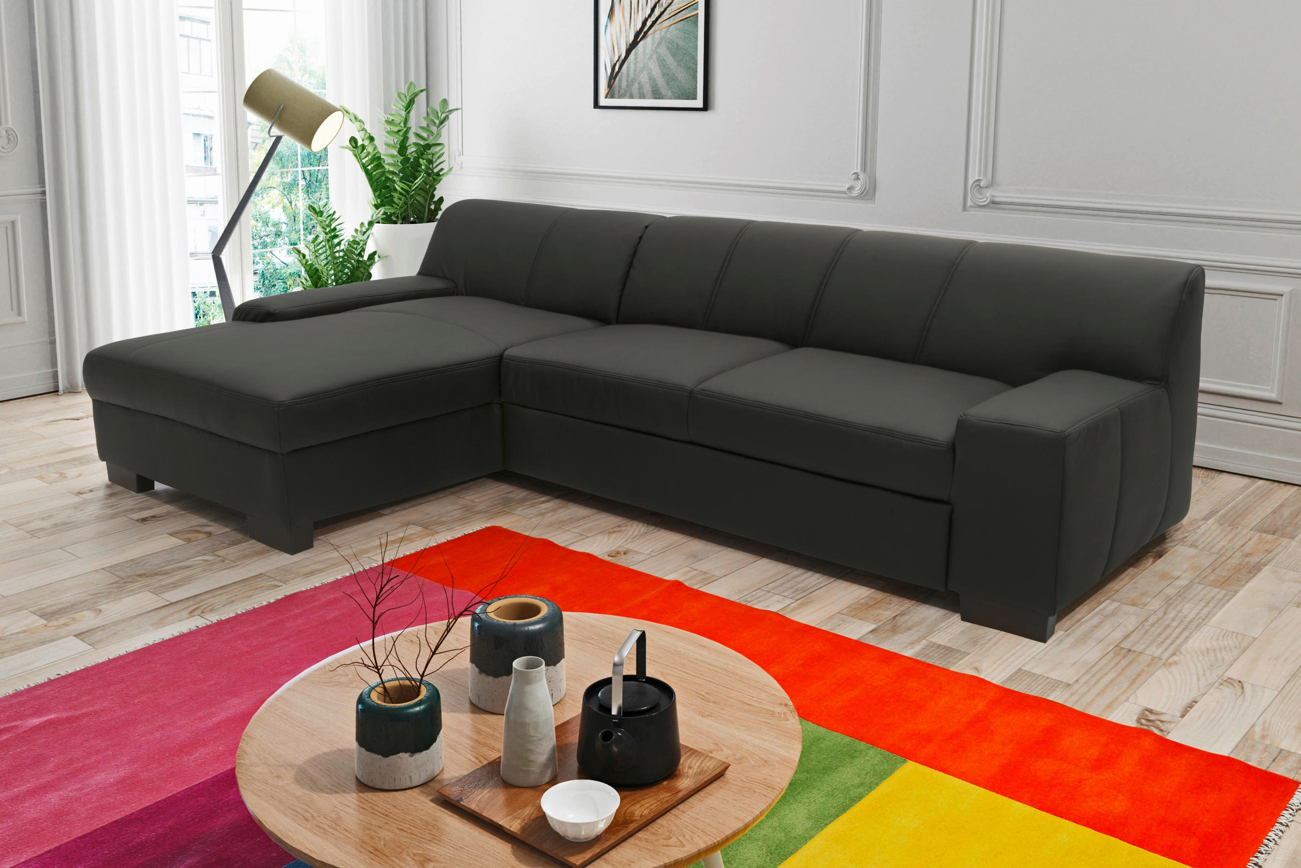 MID.YOU ECKSOFA Schwarz Lederlook, links davon: Hochflorteppich, Gardine, Topfpflanze in grün, rechts davon: Bild in hellem Wohnzimmer.