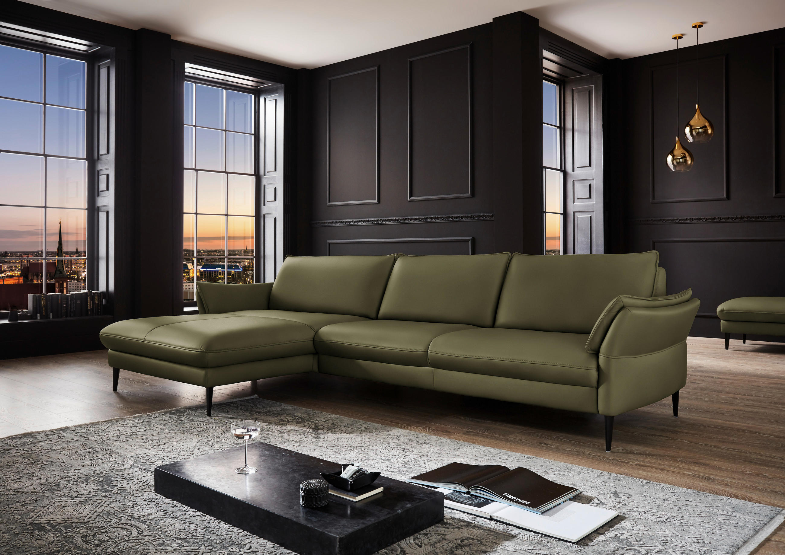 Hukla ECKSOFA Grün Echtleder, links davon: Hochflorteppich, Couchtisch in schwarz, rechts davon: Lektüre, Deckenbeleuchtung in dunklem Wohnzimmer.