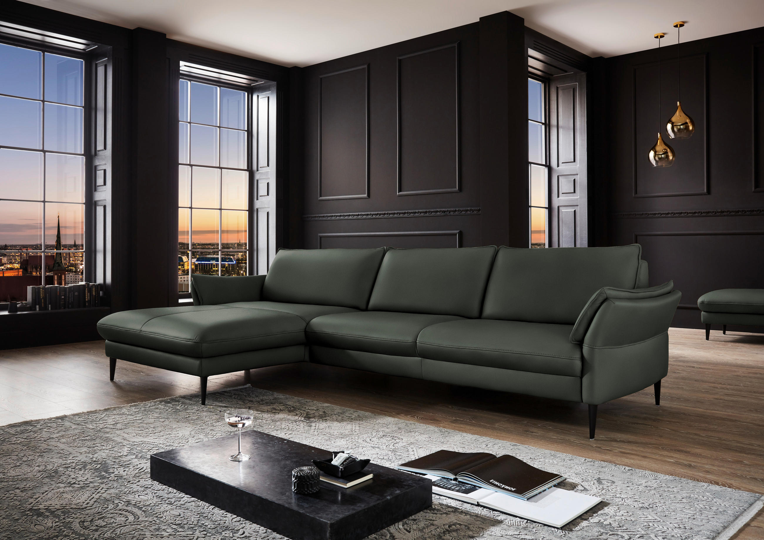 Hukla ECKSOFA inkl. Funktion Olivgrün Echtleder, links davon: Hochflorteppich in grau, Couchtisch in schwarz, rechts davon: Lektüre, Deckenbeleuchtung in dunklem Wohnzimmer.