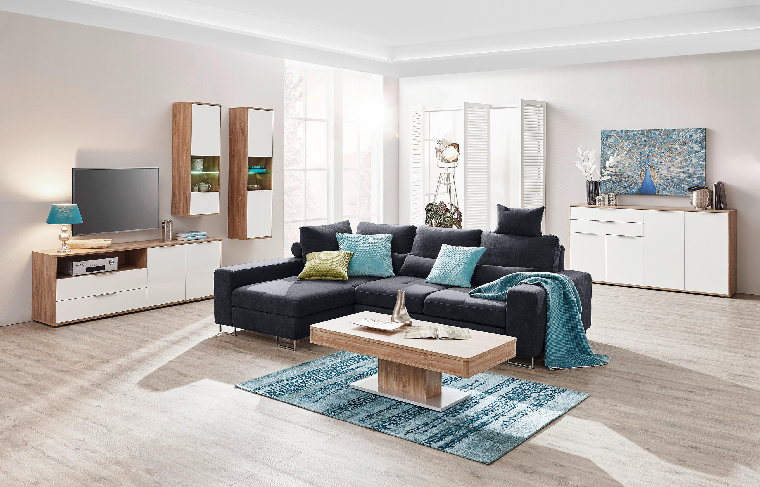 Hom`in ECKSOFA Schwarz Flachgewebe, links davon: Lowboard, rechts davon: Hochflorteppich in blau, Couchtisch, Decke in hellem Wohnzimmer.