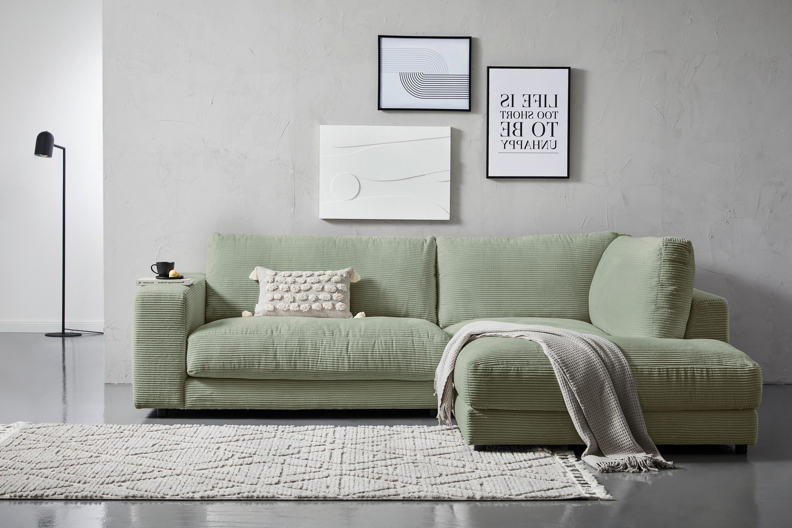 Hom`in ECKSOFA Dunkelgrün Cord, links davon: Läufer in weiß, rechts davon: Decke in grün, Bild in hellem Wohnzimmer.